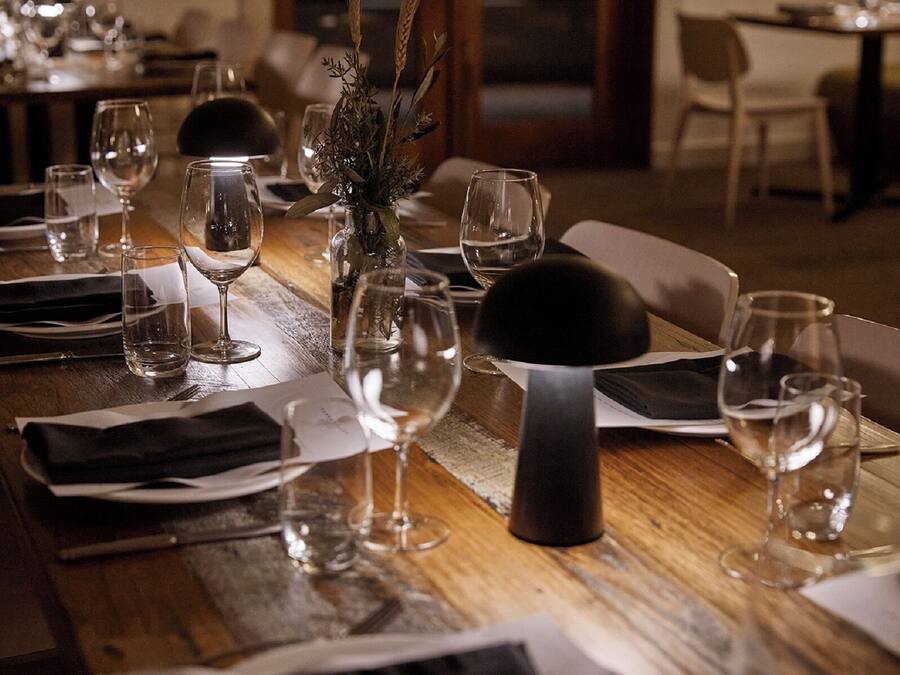 Una mesa de comedor con copas de vino, un florero con plantas secas y un sujetavasos negro.