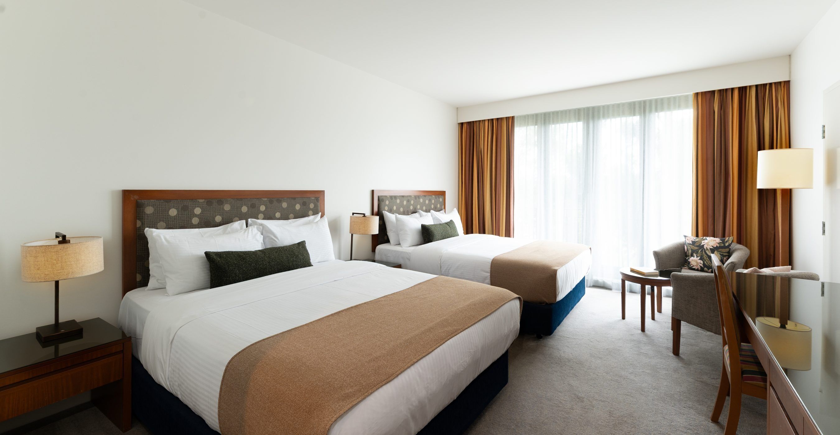Habitación de hotel con dos camas, un escritorio, una silla y un ventanal con cortinas.