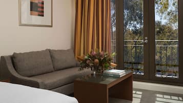 Habitación de hotel con un sofá gris, una mesita de centro con libros, una cama y un gran ventanal con cortinas.