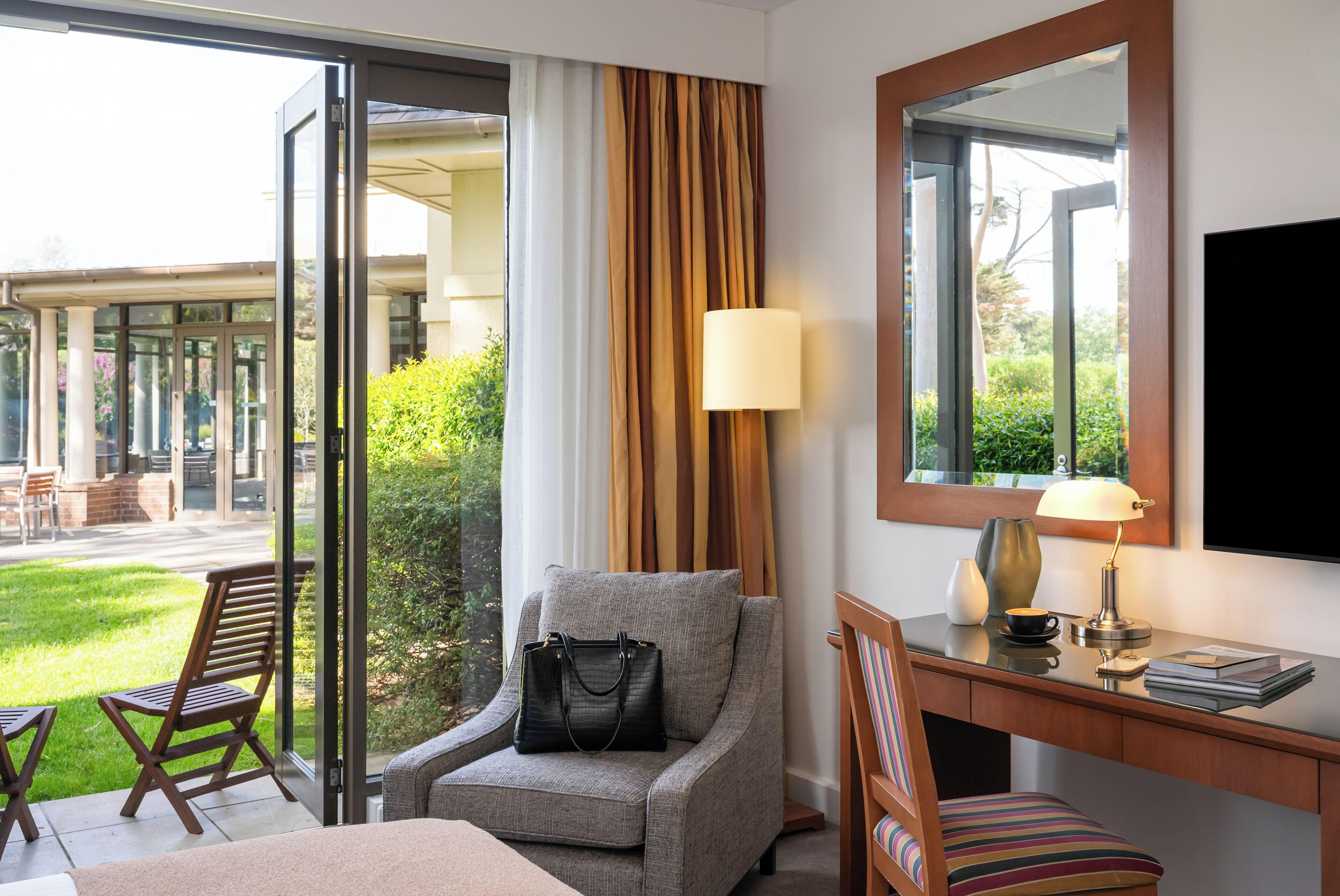 A hotel room with a sofa, a desk, a chair, a TV, a lamp, and a view of the outdoor area.