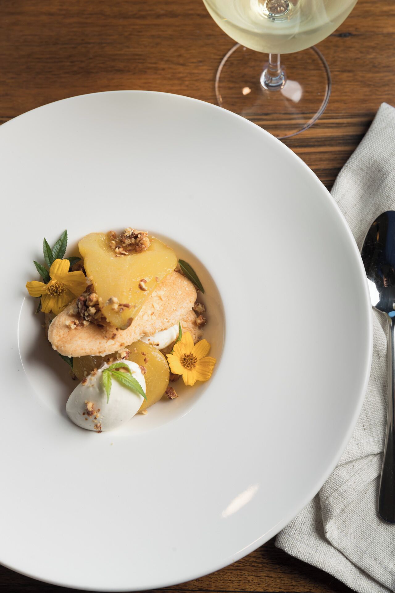 A white plate with a dessert consisting of a creamy white sphere, a slice of yellow fruit, and a nutty topping, accompanied by yellow flowers.