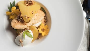A white plate with a dessert consisting of a creamy white sphere, a slice of yellow fruit, and a nutty topping, accompanied by yellow flowers.
