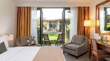 A hotel room with a bed, two armchairs, a desk, and a view of an outdoor patio with chairs and tables.