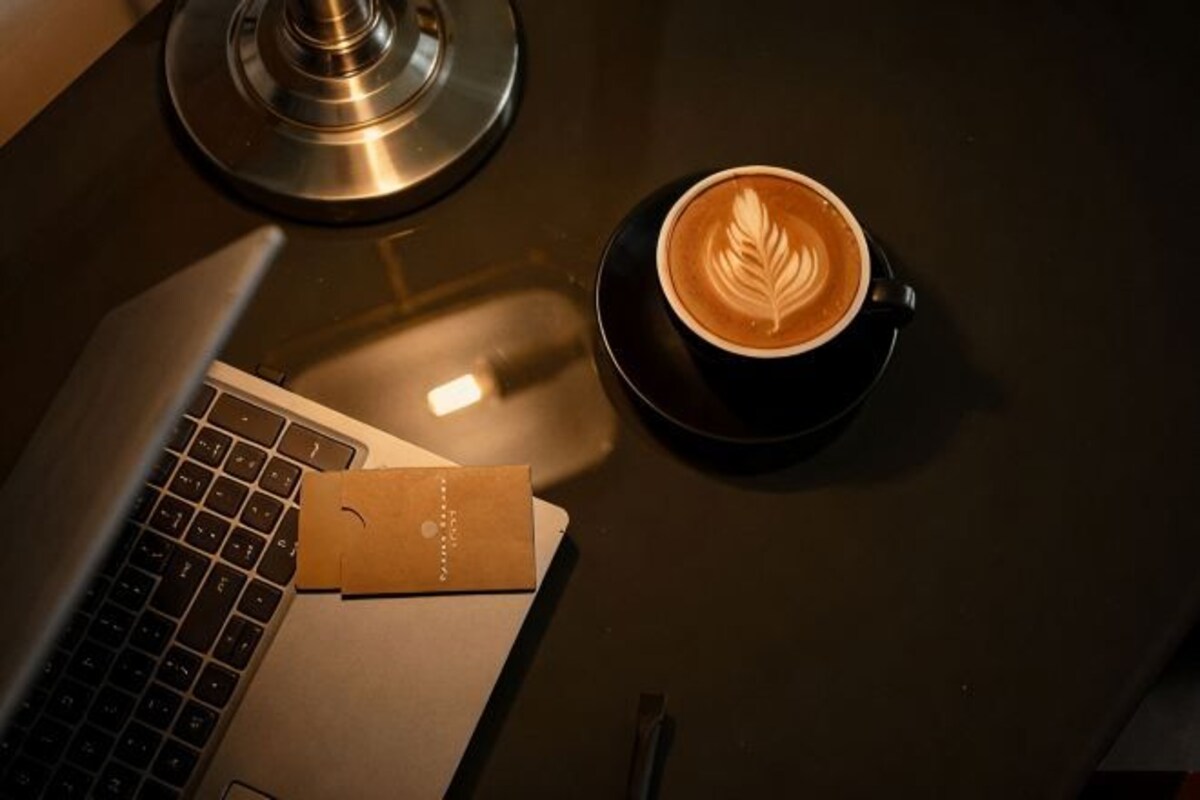 A laptop, a cup of coffee with latte art, and a coaster on a dark surface.