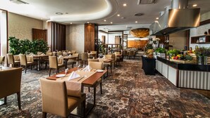Un vaste espace de restauration dans l’hôtel, avec de nombreuses tables et chaises, un bar central proposant une variété de plats, et une cuisine moderne en arrière-plan.