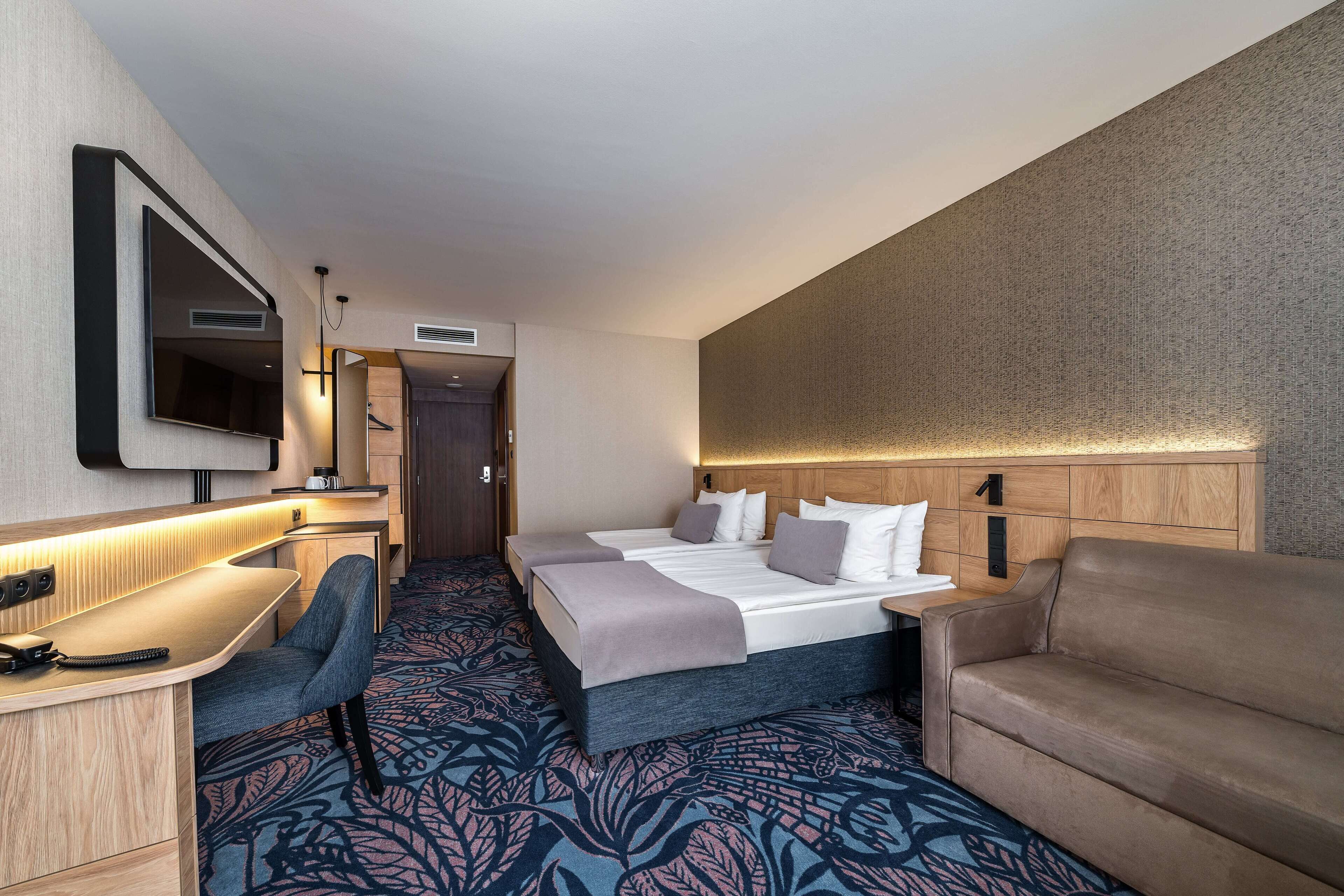 A hotel room with a bed, a desk, a chair, a TV, and a patterned carpet.