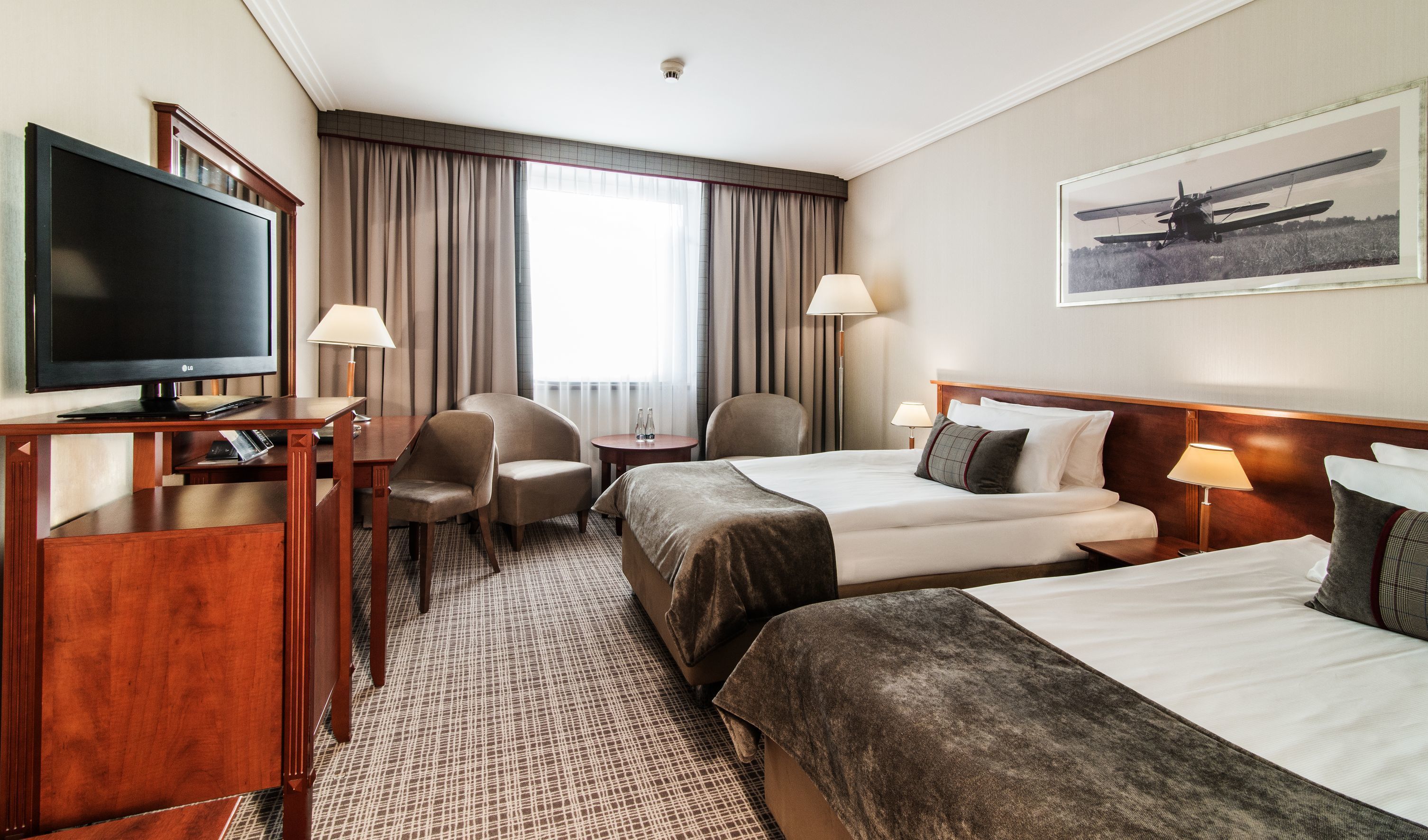 Une chambre d’hôtel avec deux lits, un bureau avec une télévision, une chaise, une petite table et une fenêtre avec des rideaux.