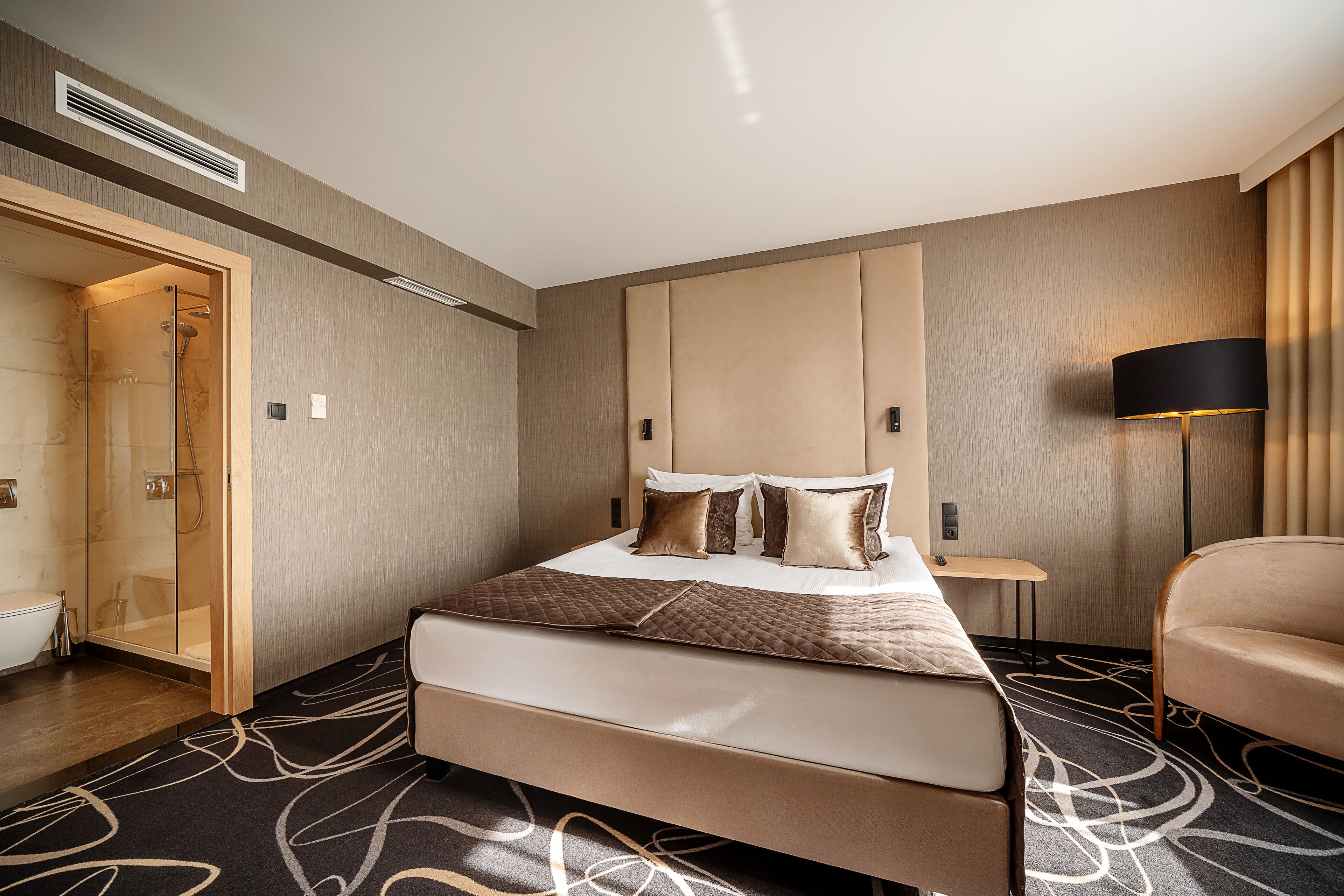 A modern hotel room with a large bed, a walk-in shower, a bedside table, and a lamp.