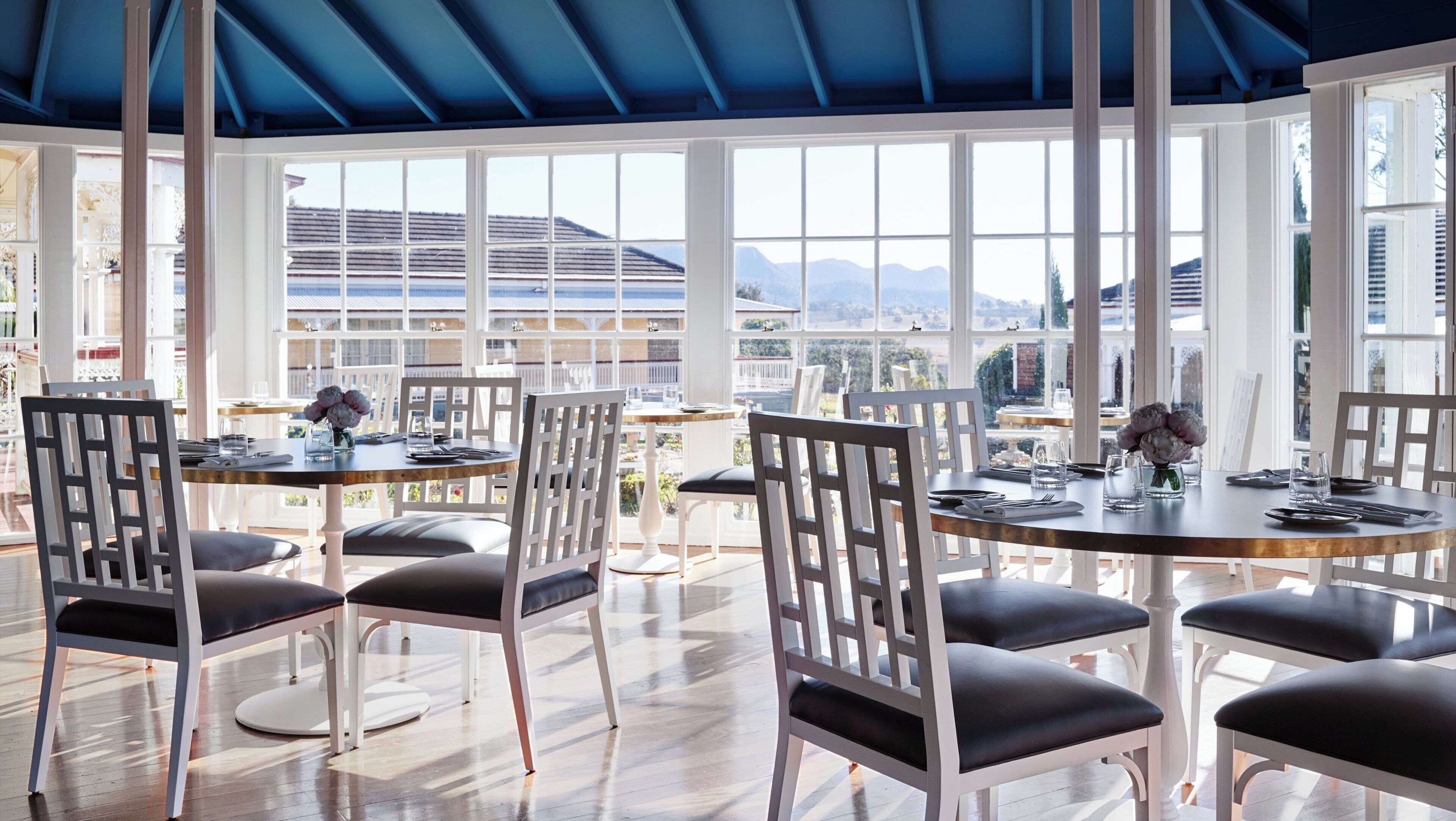 Un espace repas avec des chaises et des tables blanches, de grandes fenêtres offrant une vue sur les montagnes et un bâtiment, et un plafond bleu.