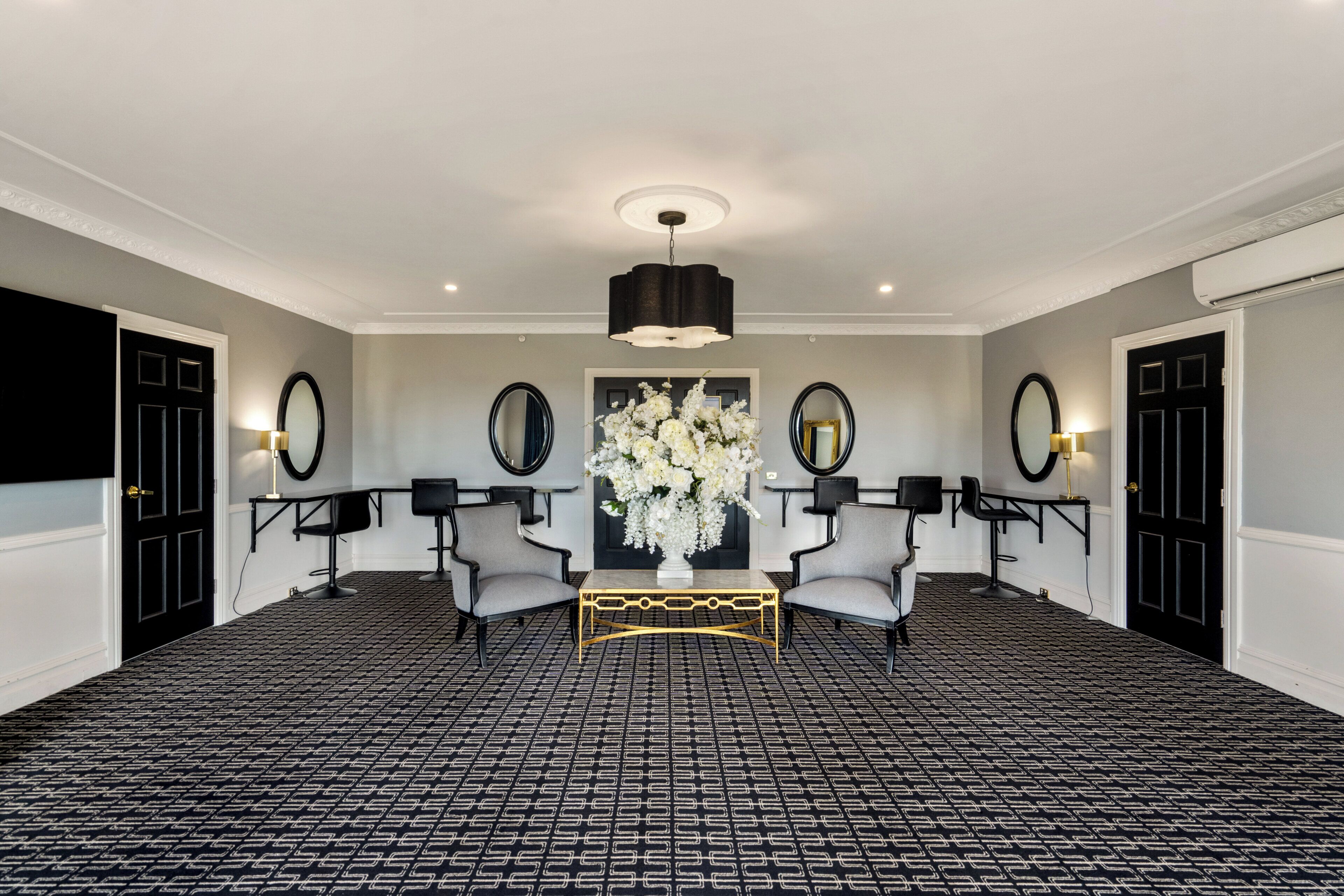 A hotel lobby with a patterned floor, two armchairs, a central table, a chandelier, and two doors.
