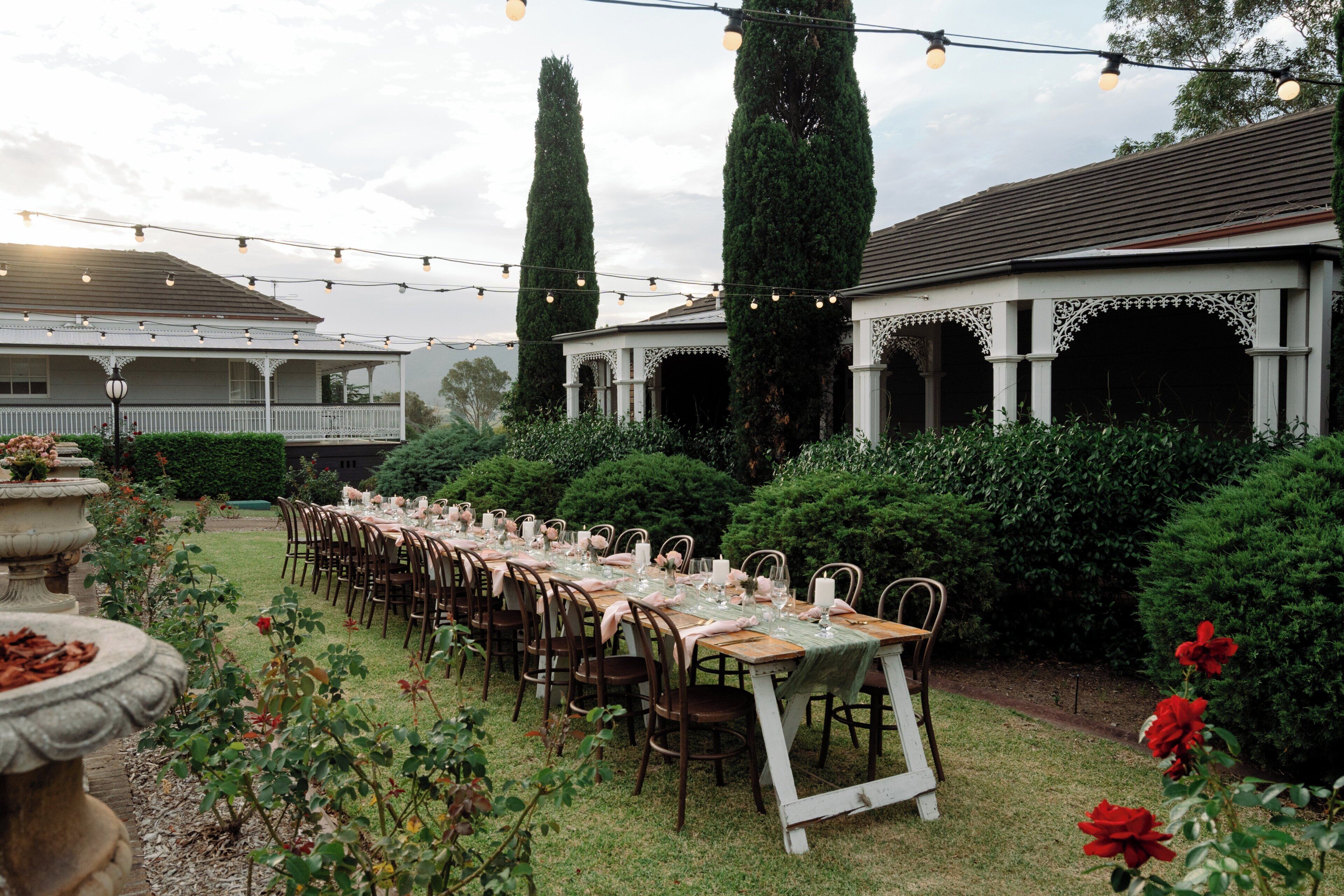 An outdoor dining area with a long table set for a meal, surrounded by greenery and a gazebo structure.