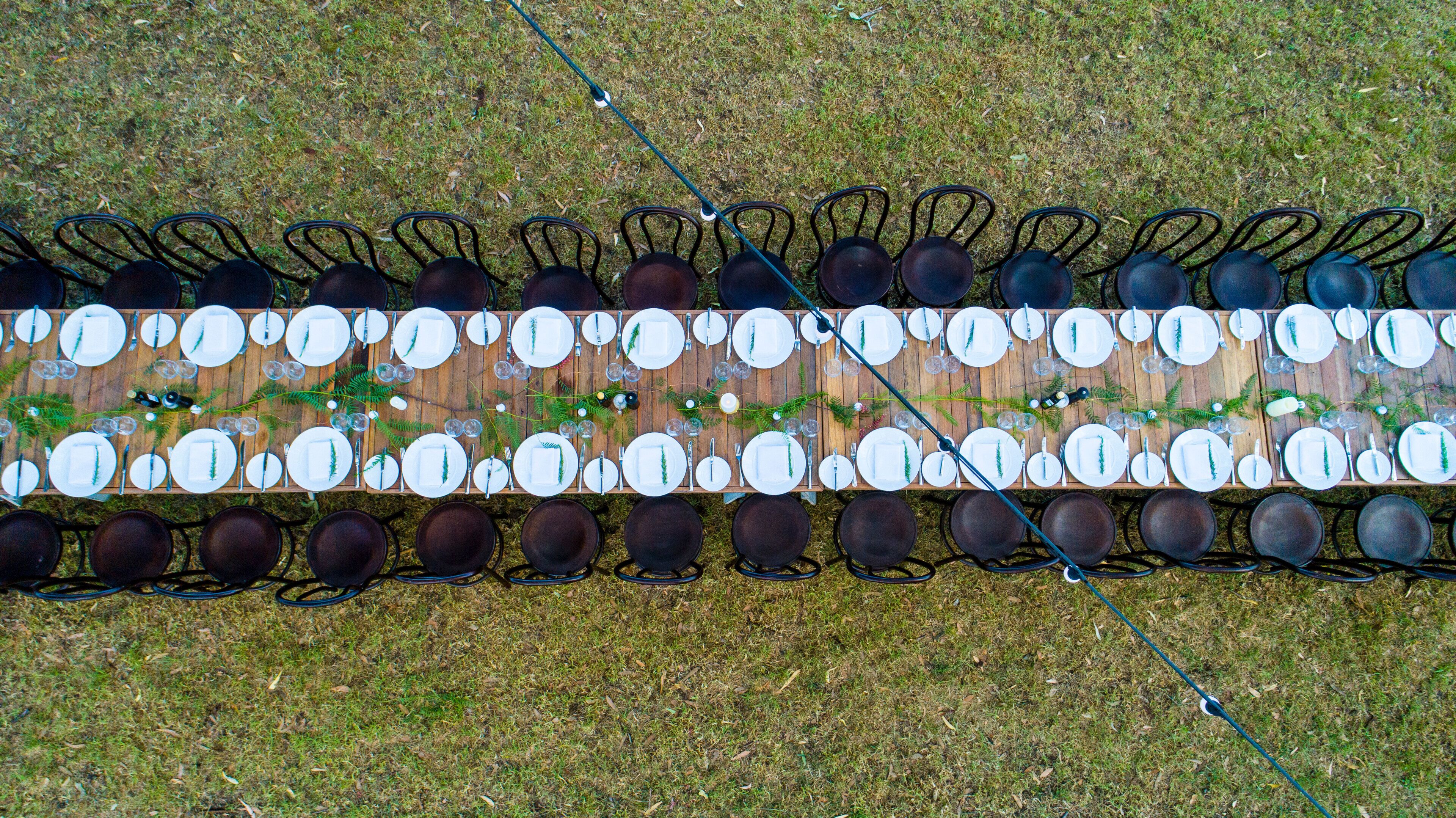A long dining table set with multiple chairs, plates, and cutlery, arranged outdoors on grass.