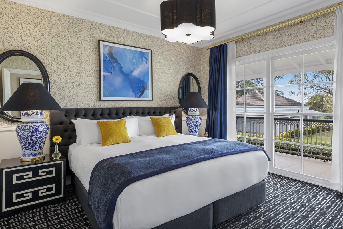 A hotel room with a large bed, two bedside lamps, a blue and white vase, a framed abstract painting, and a view of the outside.