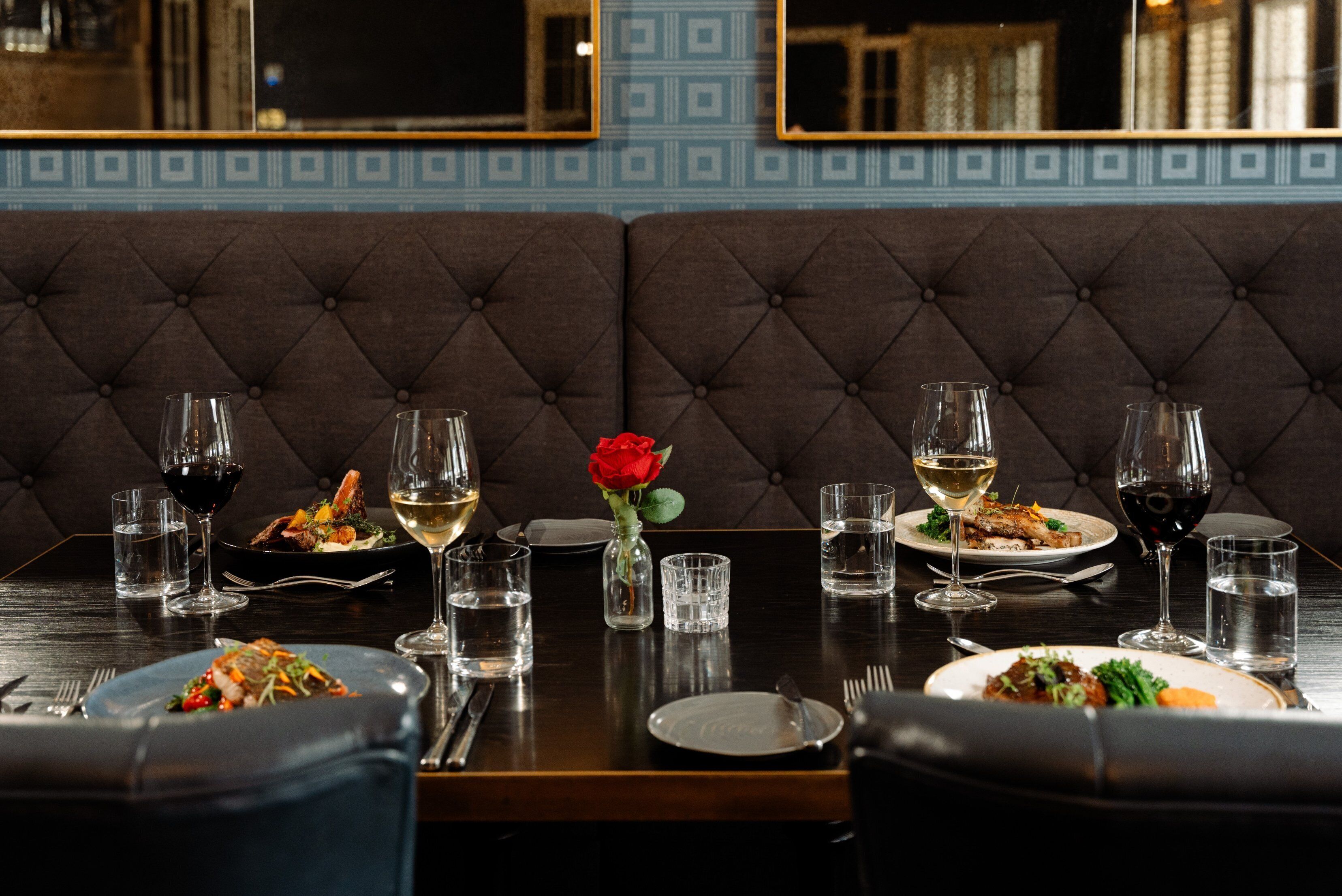 A dining table set with plates, glasses, and a vase with a rose.