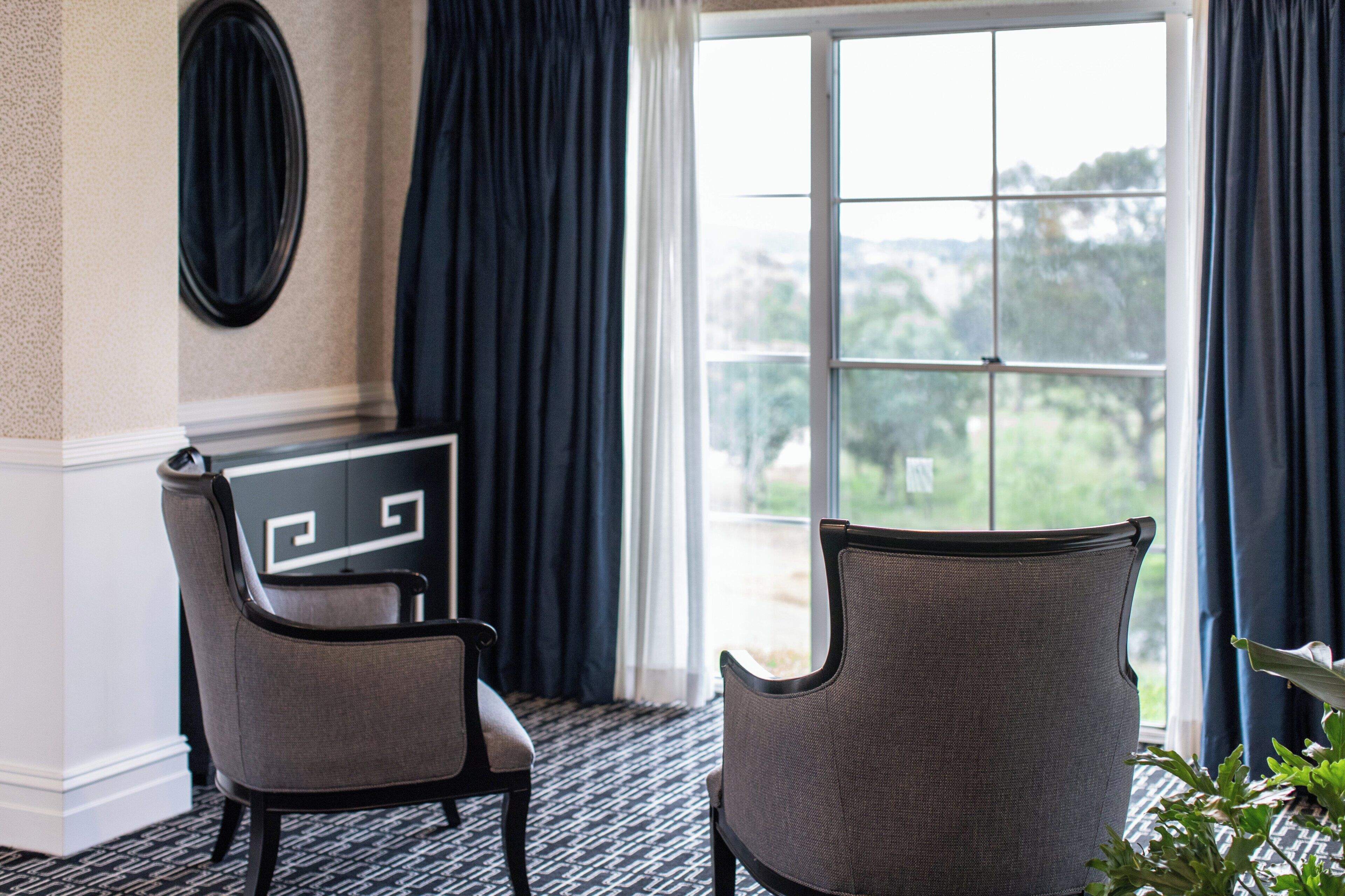 A room with two chairs, a desk, a large window with curtains, and a view of greenery outside.