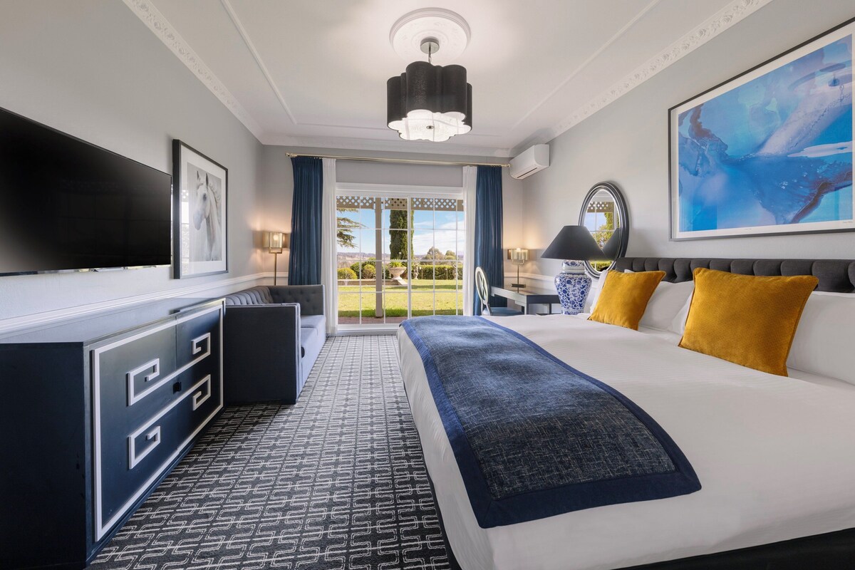 A modern hotel room with a large bed, a flat-screen TV, a desk with a chair, and a view of the outdoors through a glass door.