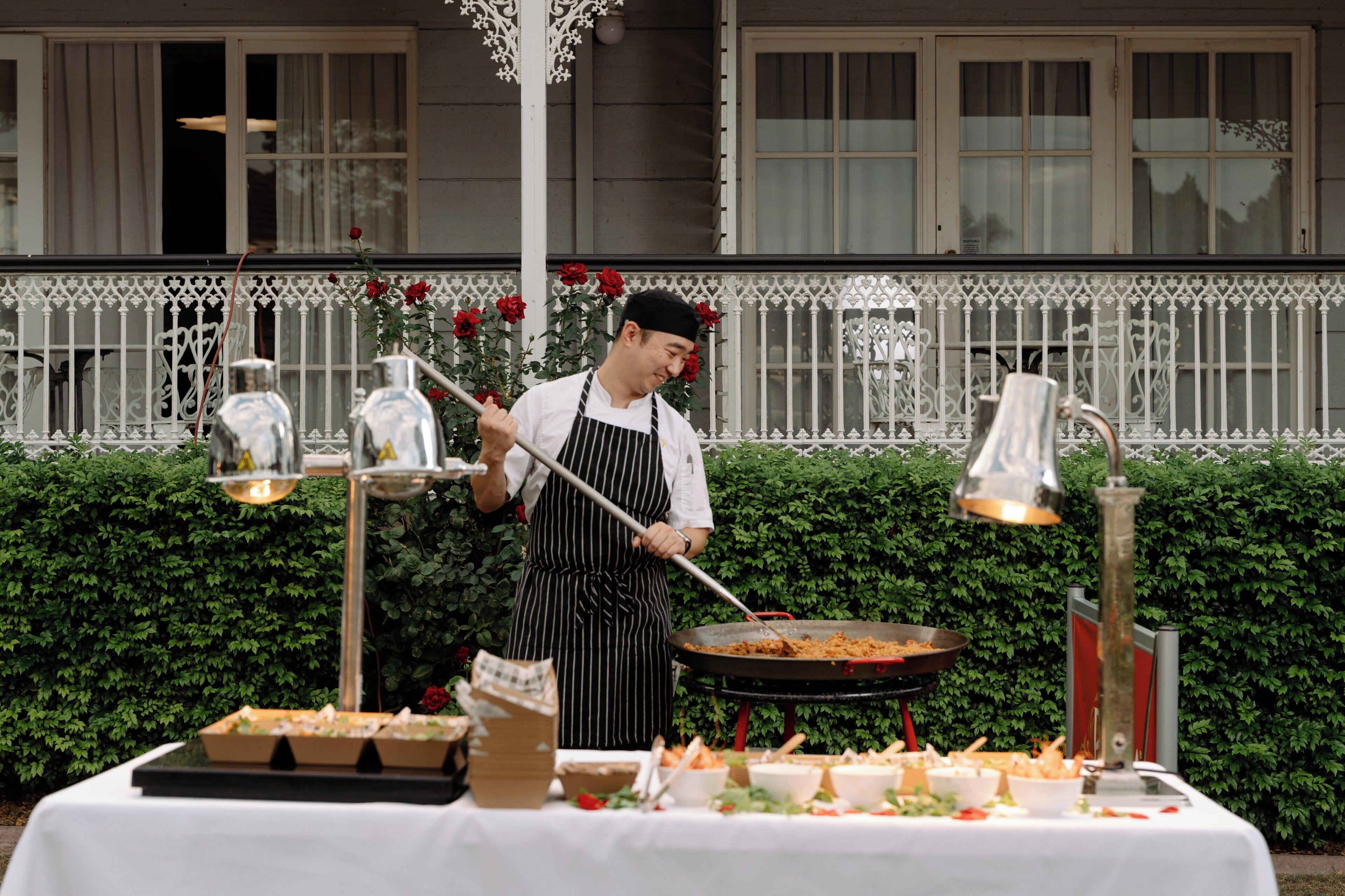 A chef cooking outdoors with a table of food in front of a white fence and green hedge.