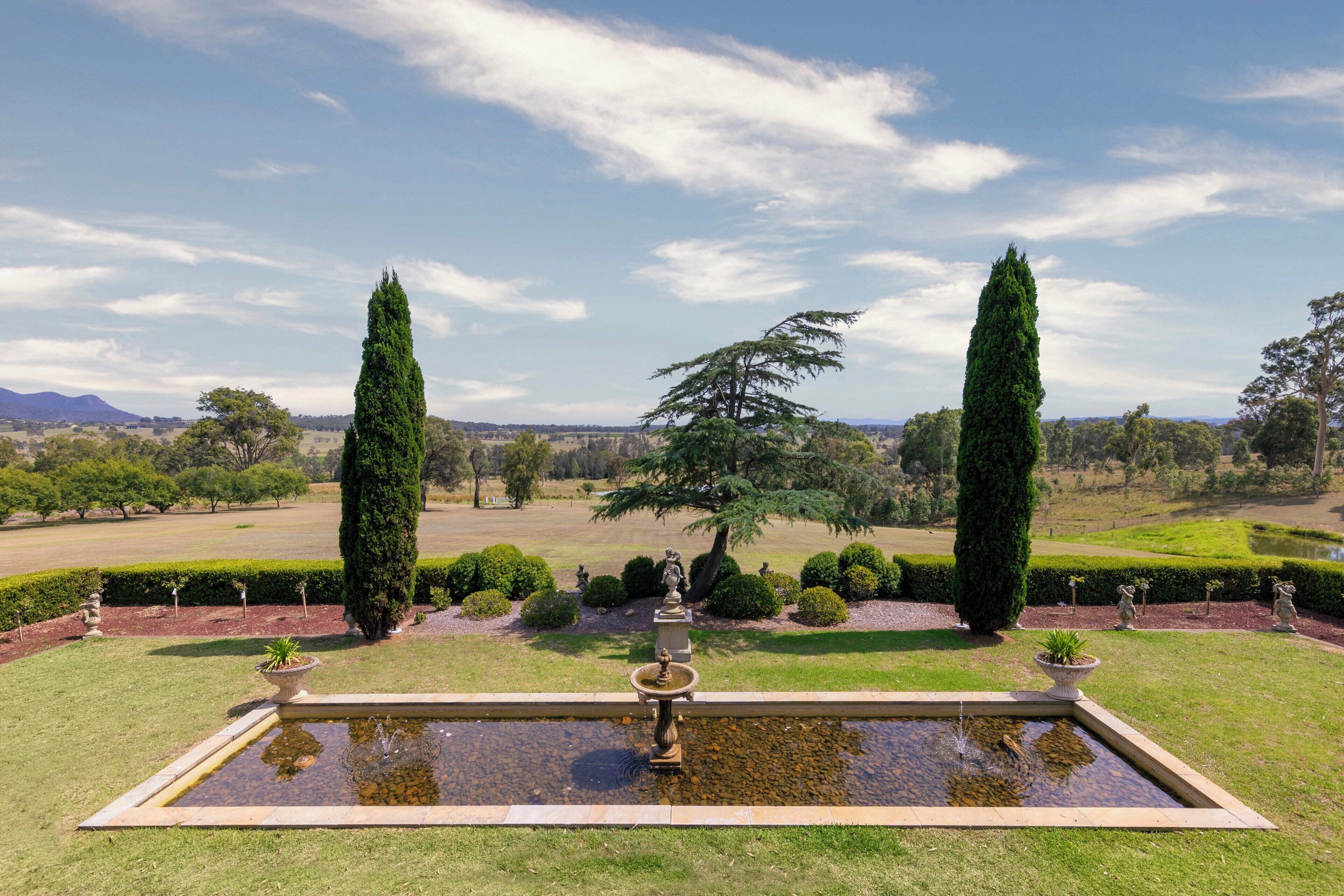 A formal garden with a central fountain, neatly trimmed hedges, and tall trees.