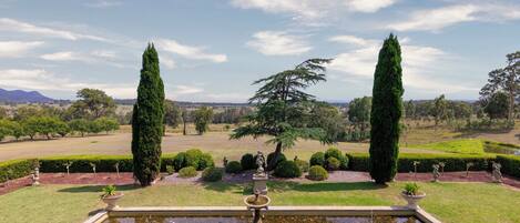 A formal garden with a central fountain, neatly trimmed hedges, and tall trees.