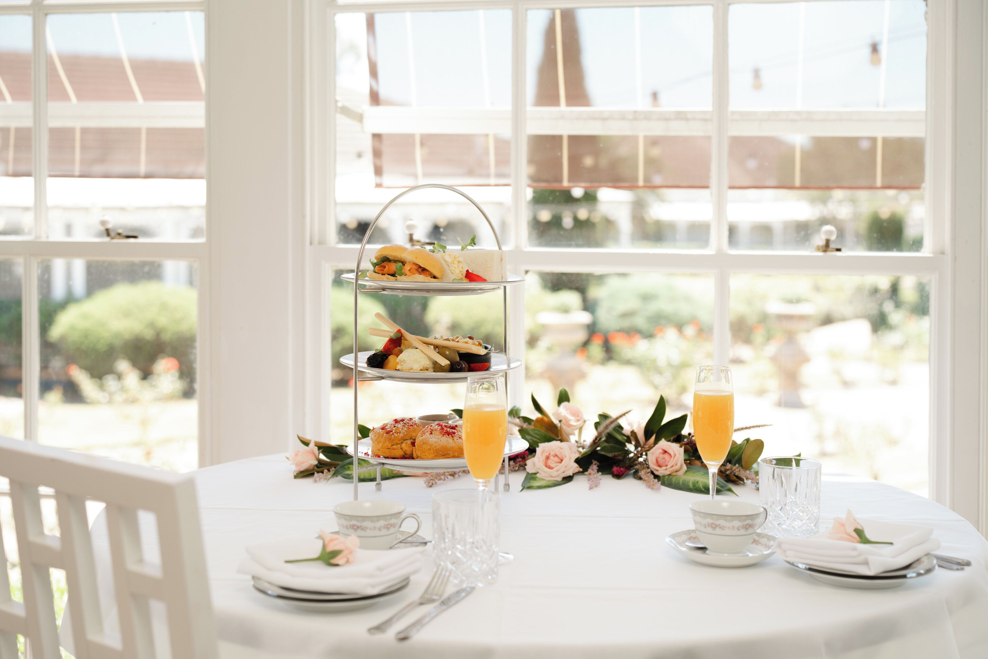 Un soporte de tres niveles para postres con pastelería y un vaso de jugo de naranja sobre una mesa con mantel blanco, rodeado de arreglos florales y cubiertos.