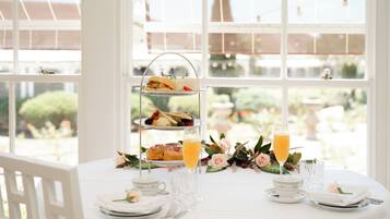 Un soporte de tres niveles para postres con pastelería y un vaso de jugo de naranja sobre una mesa con mantel blanco, rodeado de arreglos florales y cubiertos.