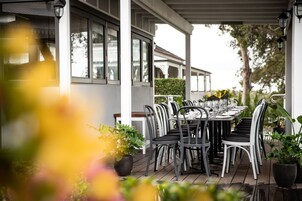 Un área de comedor al aire libre con un porche cubierto, mesas dispuestas con sillas y plantas en macetas.