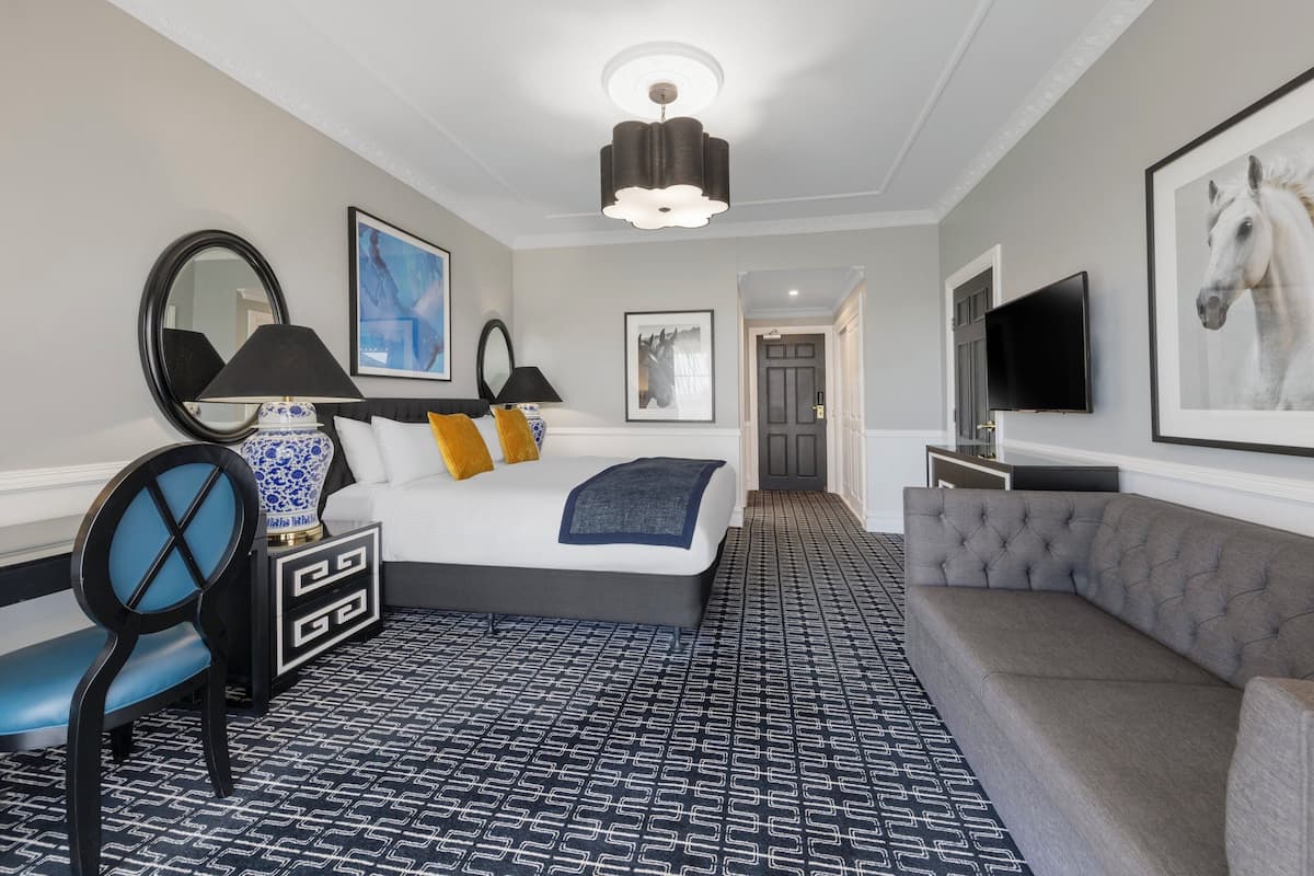Et moderne hotelværelse med en stor seng, en grå sofa, et skrivebord med lampe og billedkunst på væggene.