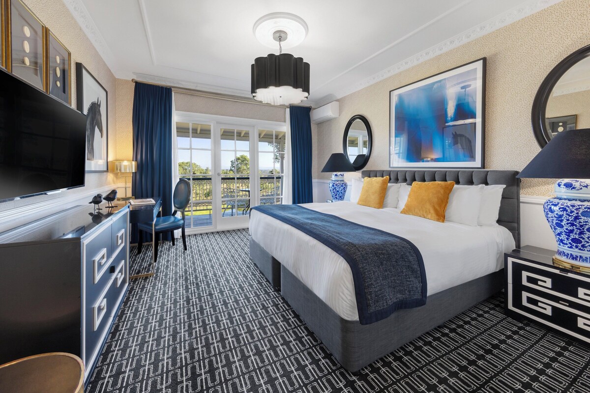 A hotel room with a large bed, a desk with a flat-screen TV, a chair, a desk lamp, and a large window with curtains.