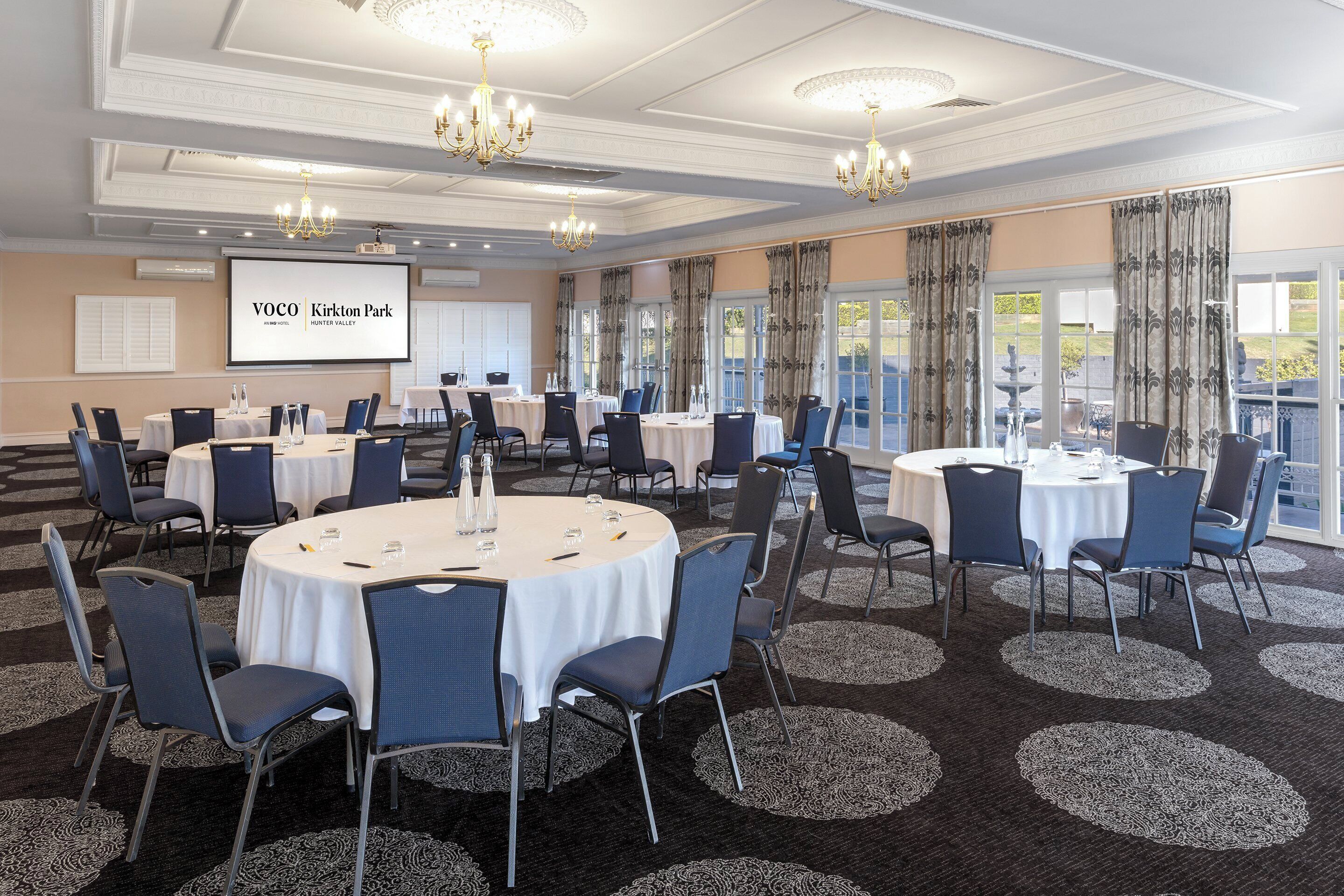 A conference room with round tables, chairs, and a projector screen displaying 'VOCO Kirkton Park'.