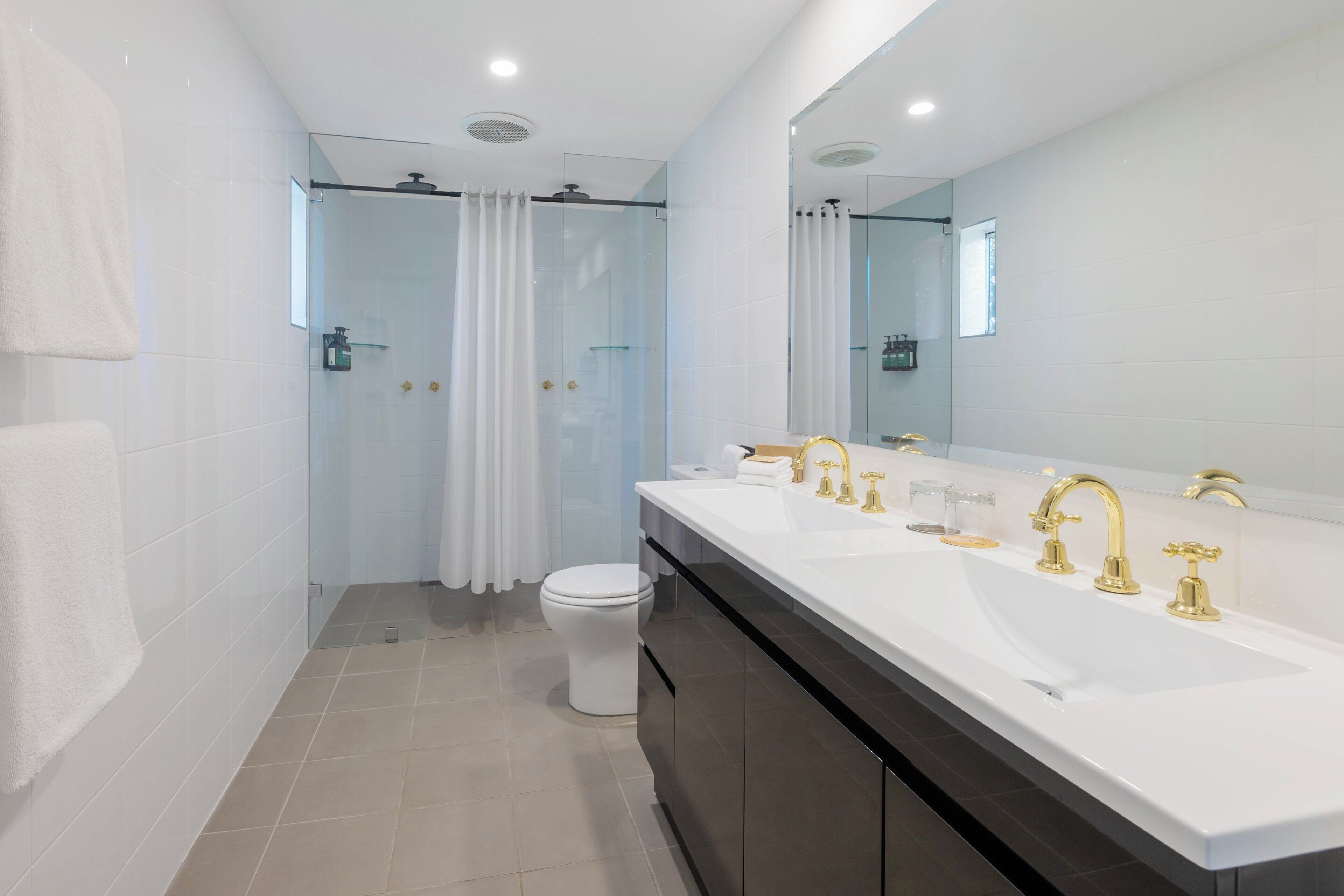 A modern bathroom with a large mirror, a double sink with gold faucets, and a glass shower enclosure.