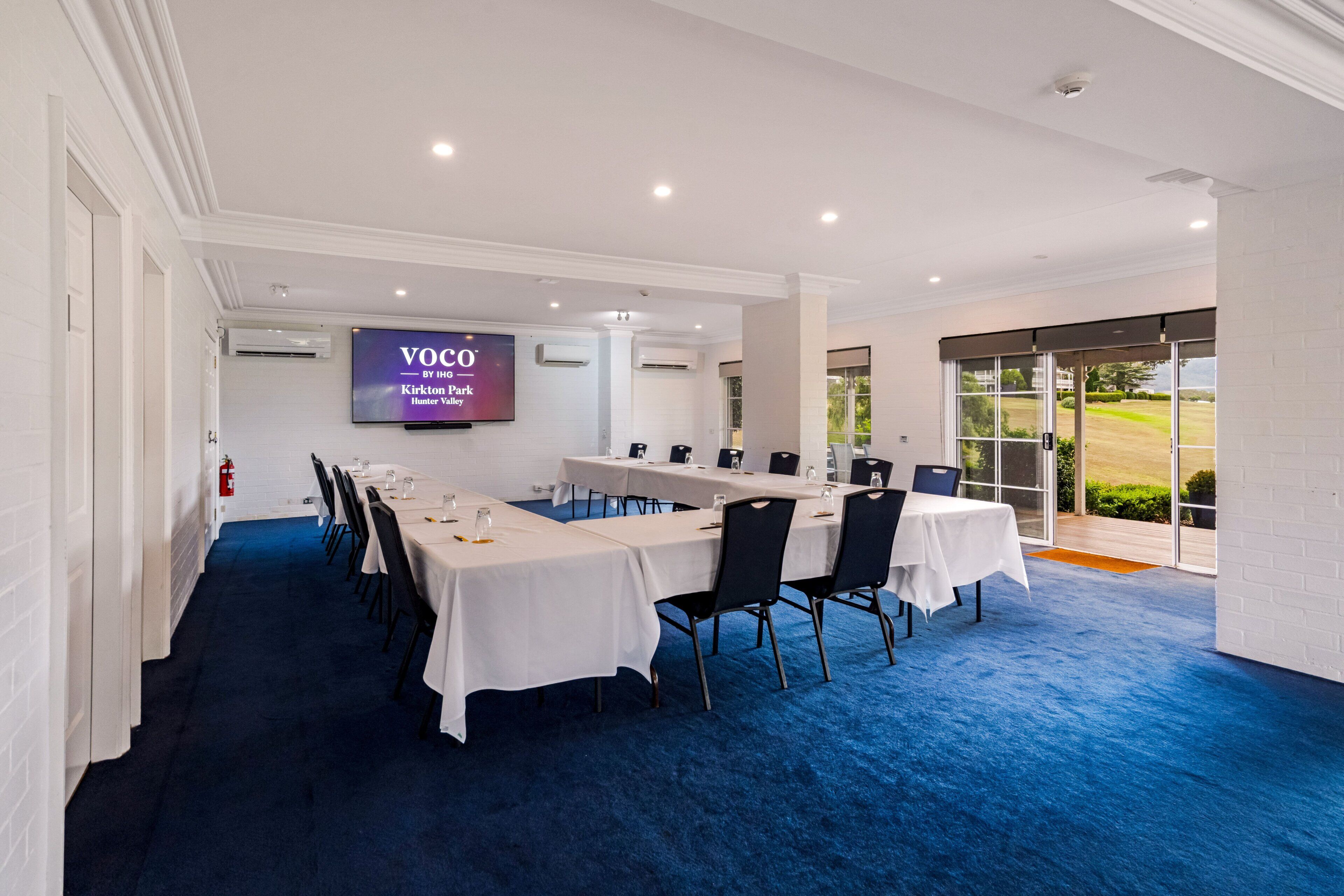 A conference room with a large screen, long tables, and chairs, with a view of a park through the windows.
