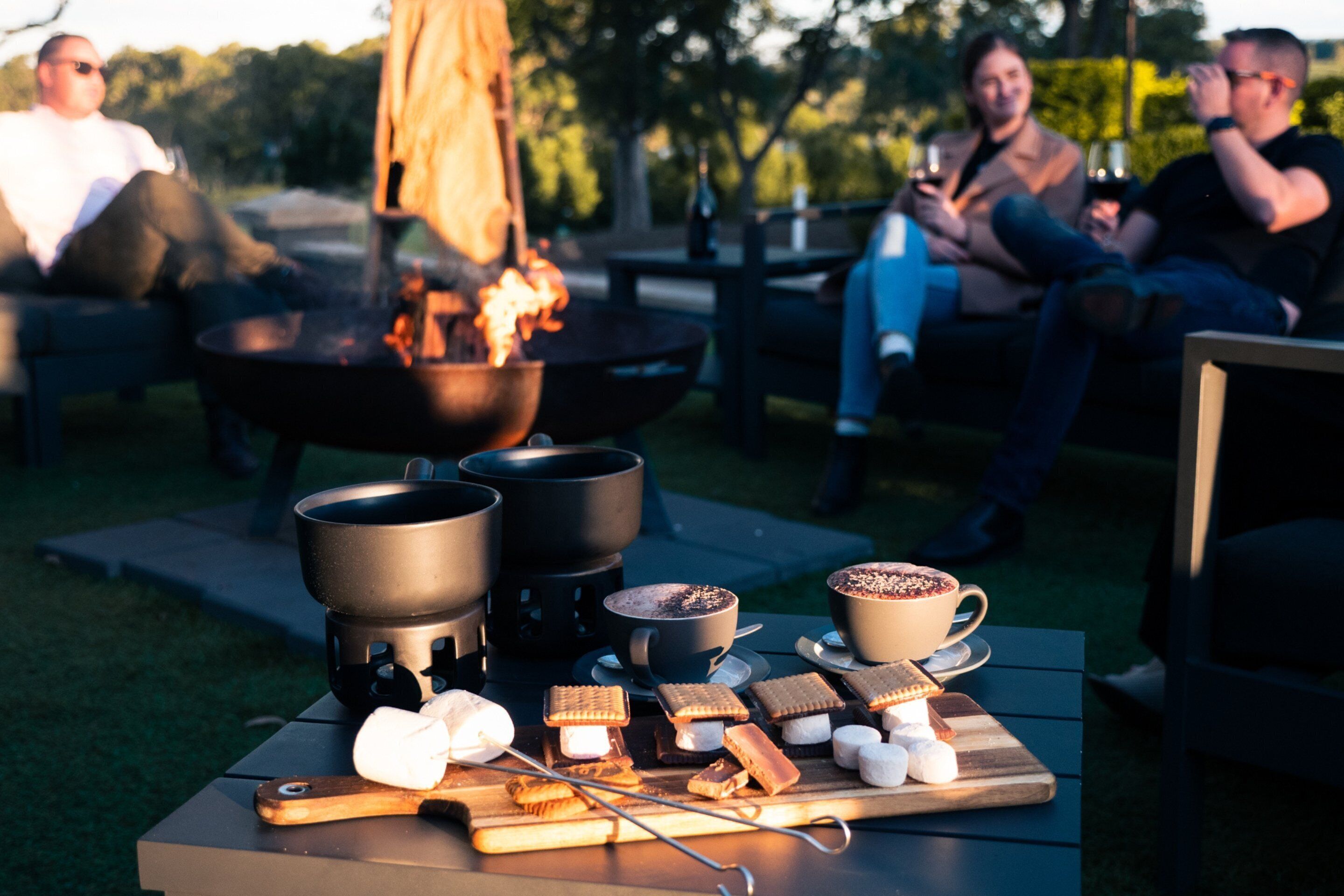 Een groep mensen zit buiten bij een vuurkuil, met marshmallows en warme drankjes op een tafel.