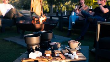 A group of people sitting outdoors with a fire pit, marshmallows, and hot beverages on a table.