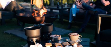 A group of people sitting outdoors with a fire pit, marshmallows, and hot beverages on a table.