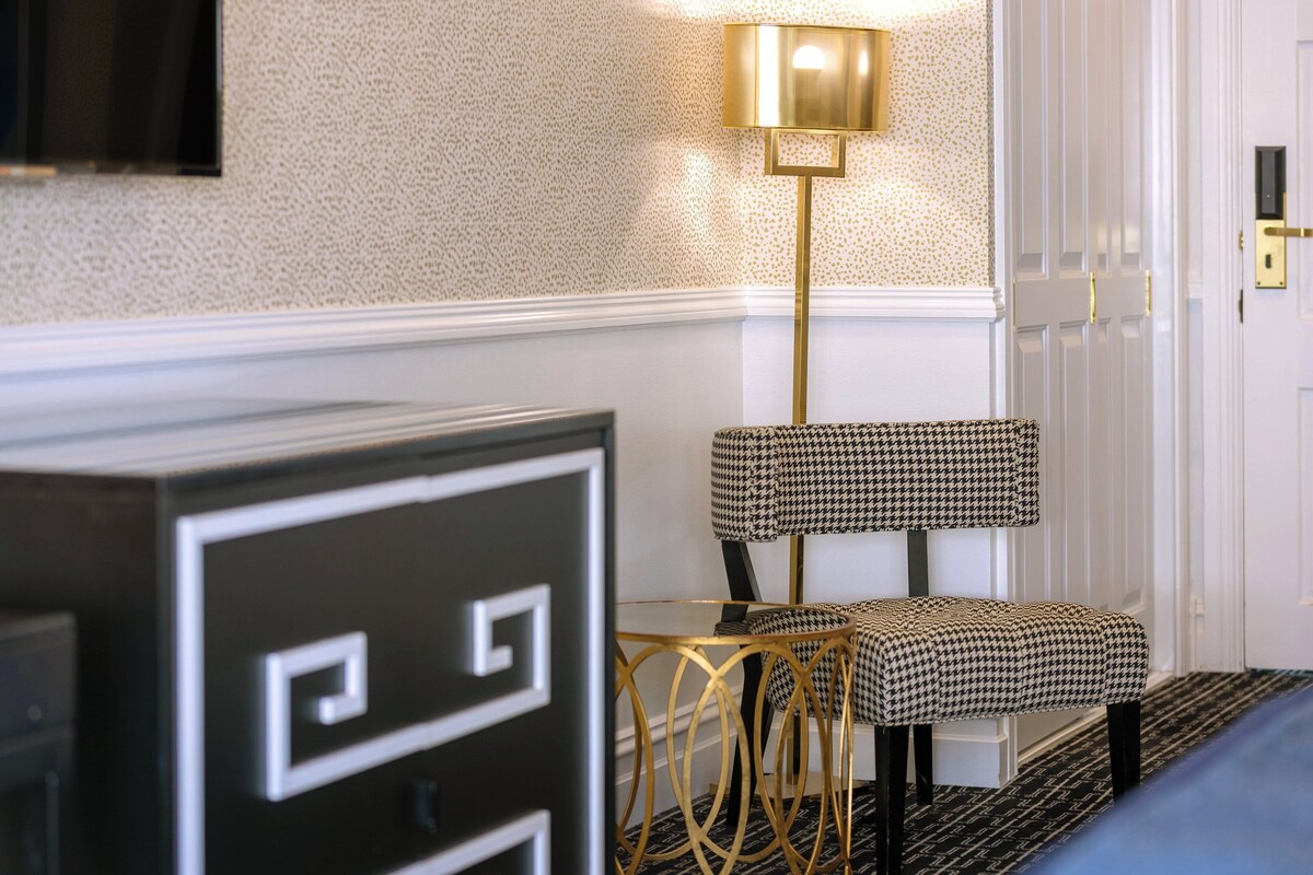 A hallway with a houndstooth patterned chair, a gold floor lamp, and a black and white cabinet.