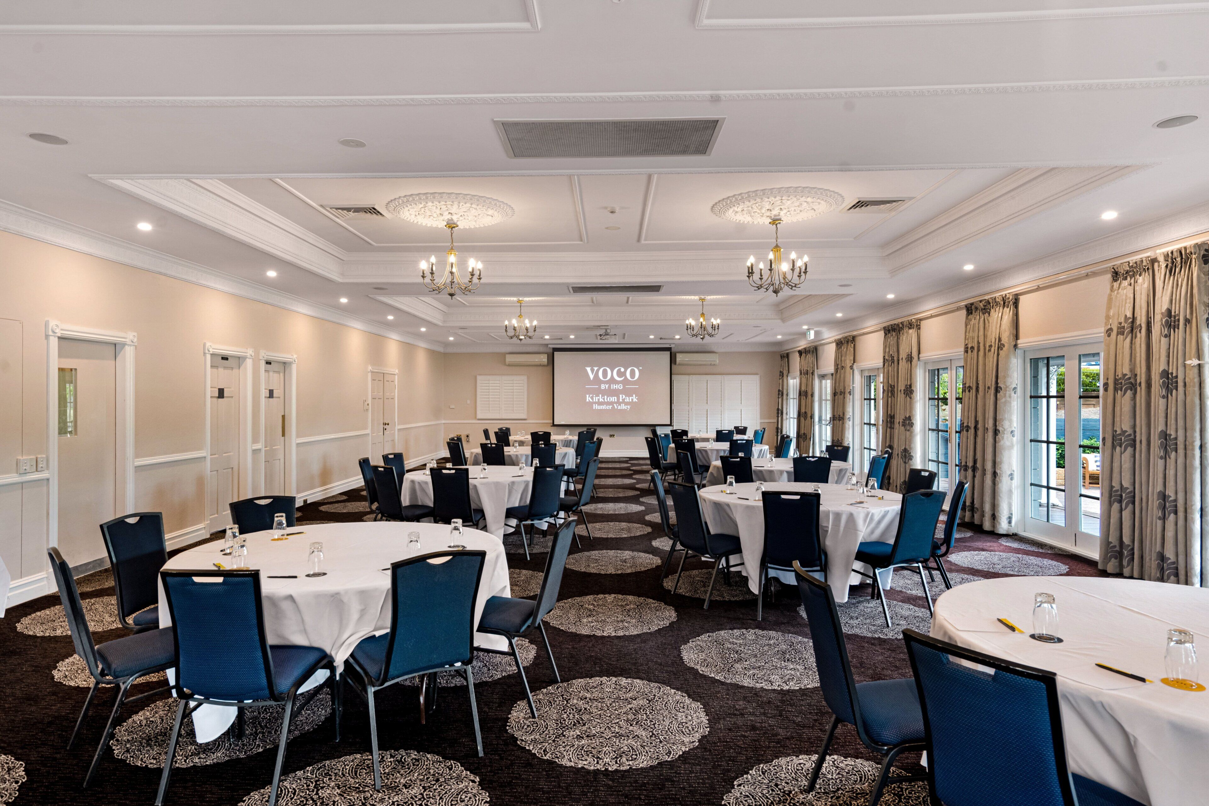 Un salón de conferencias con mesas redondas, sillas azules y una pantalla proyectora que muestra 'VOCO by Kirkton Park Hunter Valley'.