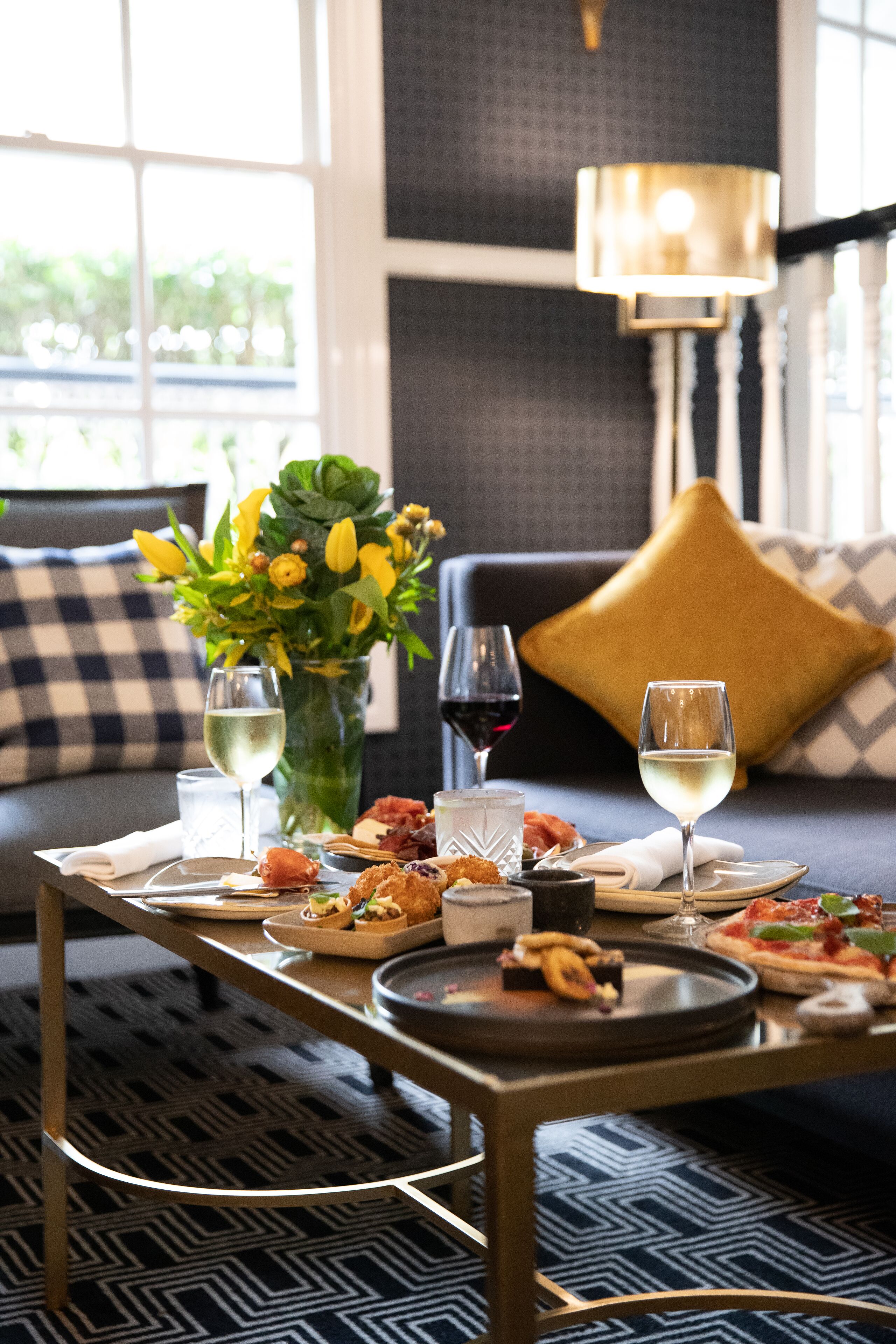 A dining table with a variety of appetizers and a bottle of wine, set on a patterned floor with a sofa and decorative pillows in the background.