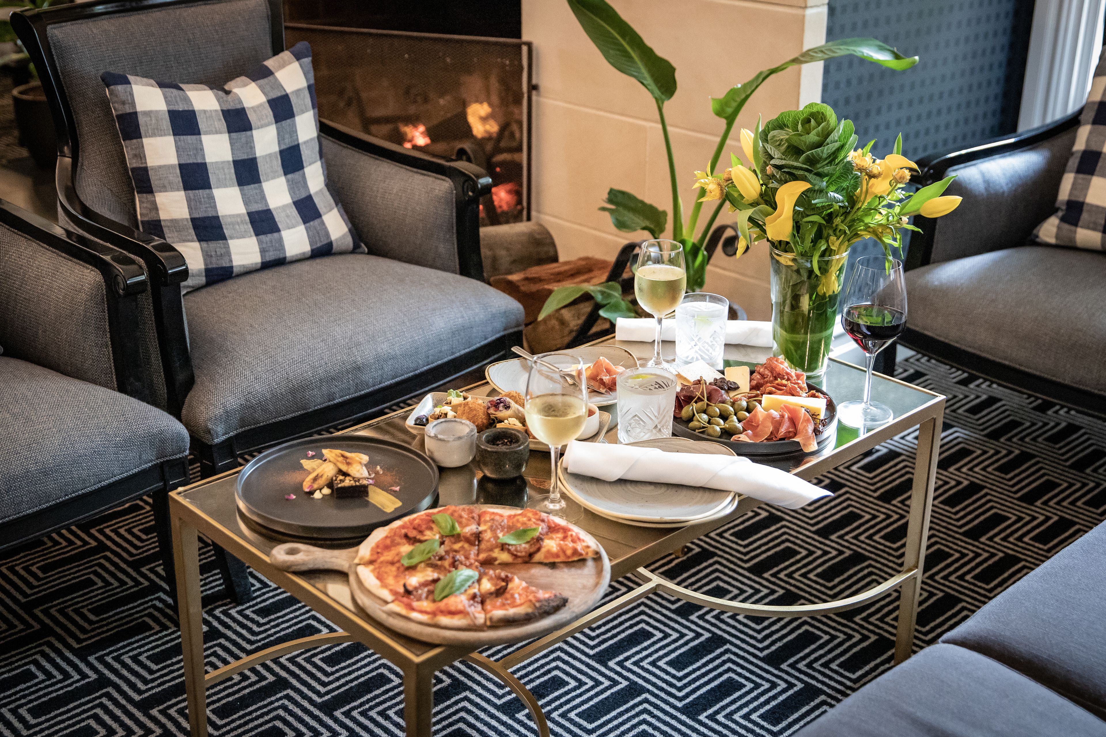 Un rincón acogedor con una chimenea, una pizza en una bandeja y una mesa con bebidas y aperitivos.