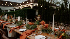 A long dining table set outdoors with candles, glassware, and greenery.