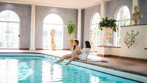 A swimming pool with two people sitting on the edge, large windows, and classical statues.