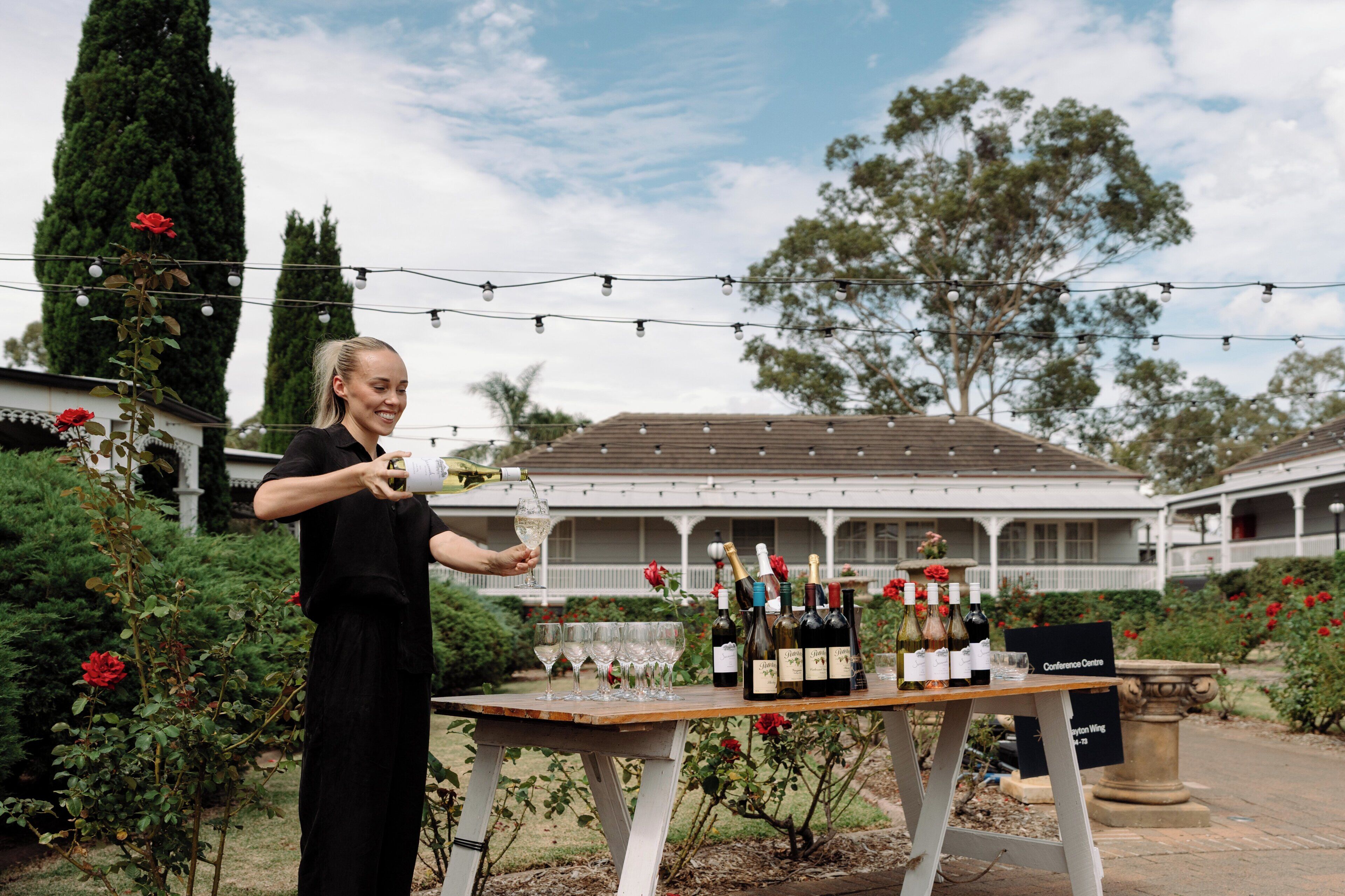 A woman pouring wine at an outdoor table with bottles and glasses, surrounded by greenery and a building with a balcony.