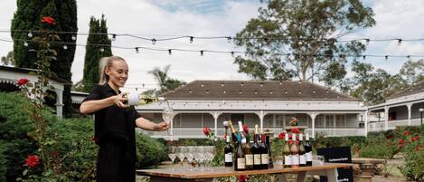 A woman pouring wine at an outdoor table with bottles and glasses, surrounded by greenery and a building with a balcony.