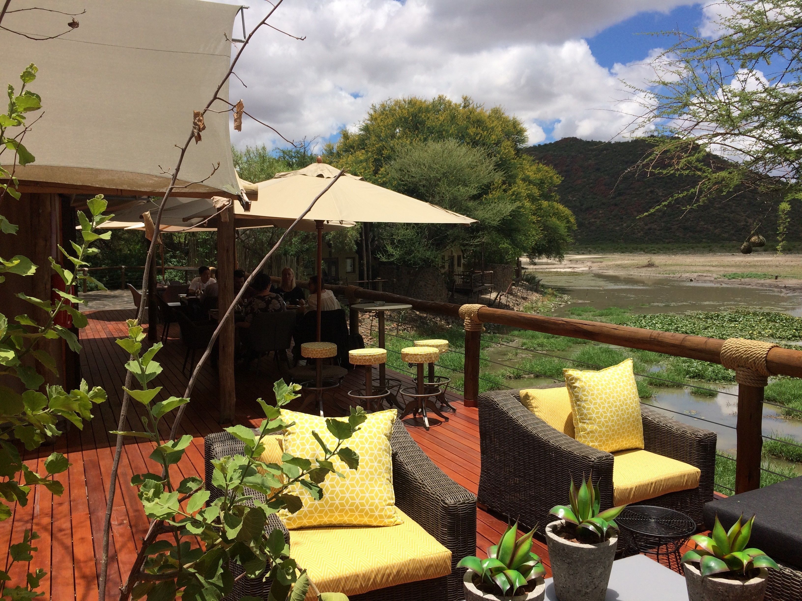 An outdoor seating area with wicker furniture and yellow cushions, overlooking a river and surrounded by greenery.