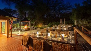 An outdoor dining area with tables set for two, surrounded by wooden deck and trees. - Tau Game Lodge (Ramotshere Moiloa)