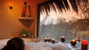 A spa setting with a hot tub filled with bubbles, candles, and a view of trees through a window. - Tau Game Lodge (Ramotshere Moiloa)