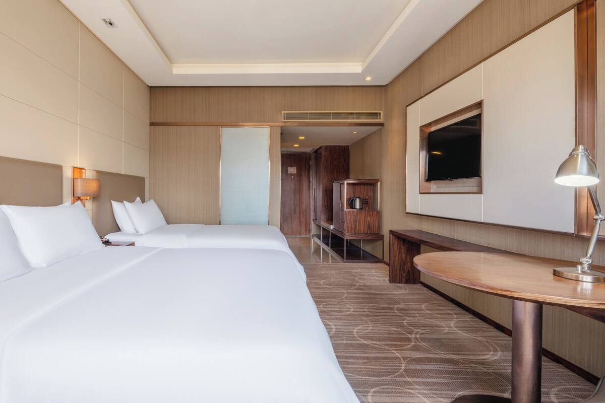 Habitación de hotel con dos camas, un escritorio, un televisor y un baño visible a través de una puerta.