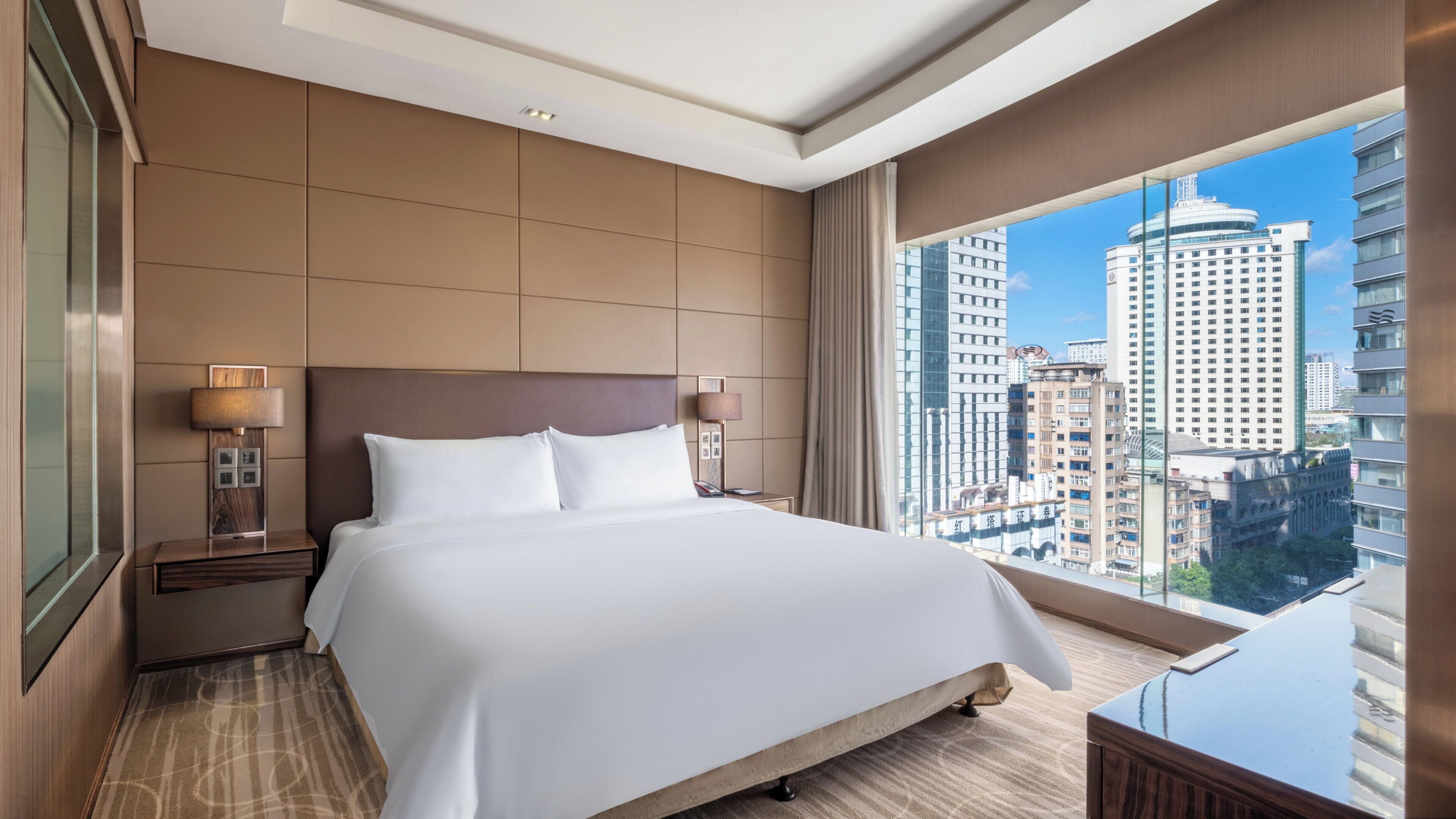 Ett hotellrum med en stor säng, ett nattduksbord med en lampa, utsikt över stadsbilden genom fönstret och en modern design.