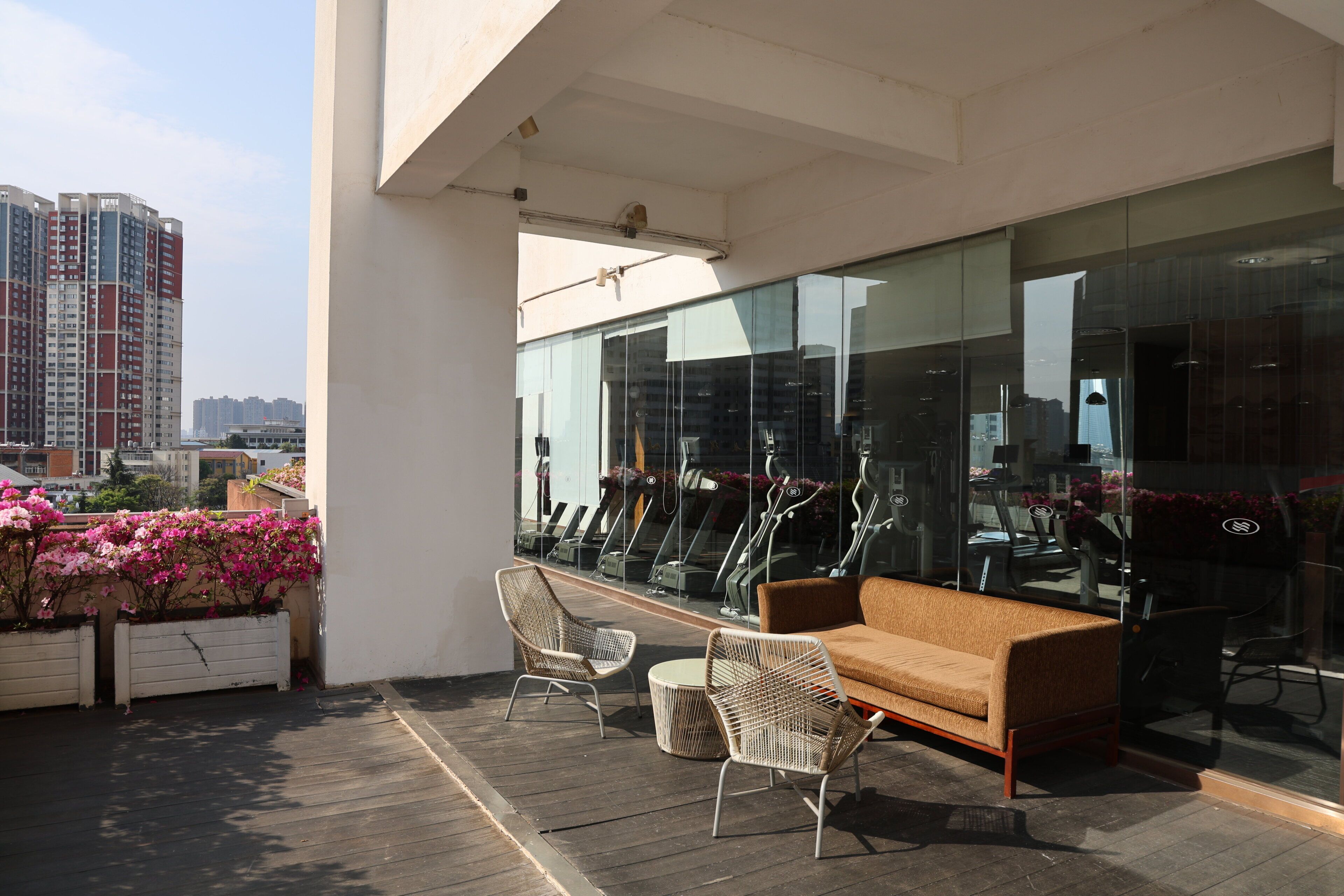 A modern outdoor seating area with a sofa and chairs, a glass wall reflecting gym equipment, and a view of high-rise buildings.