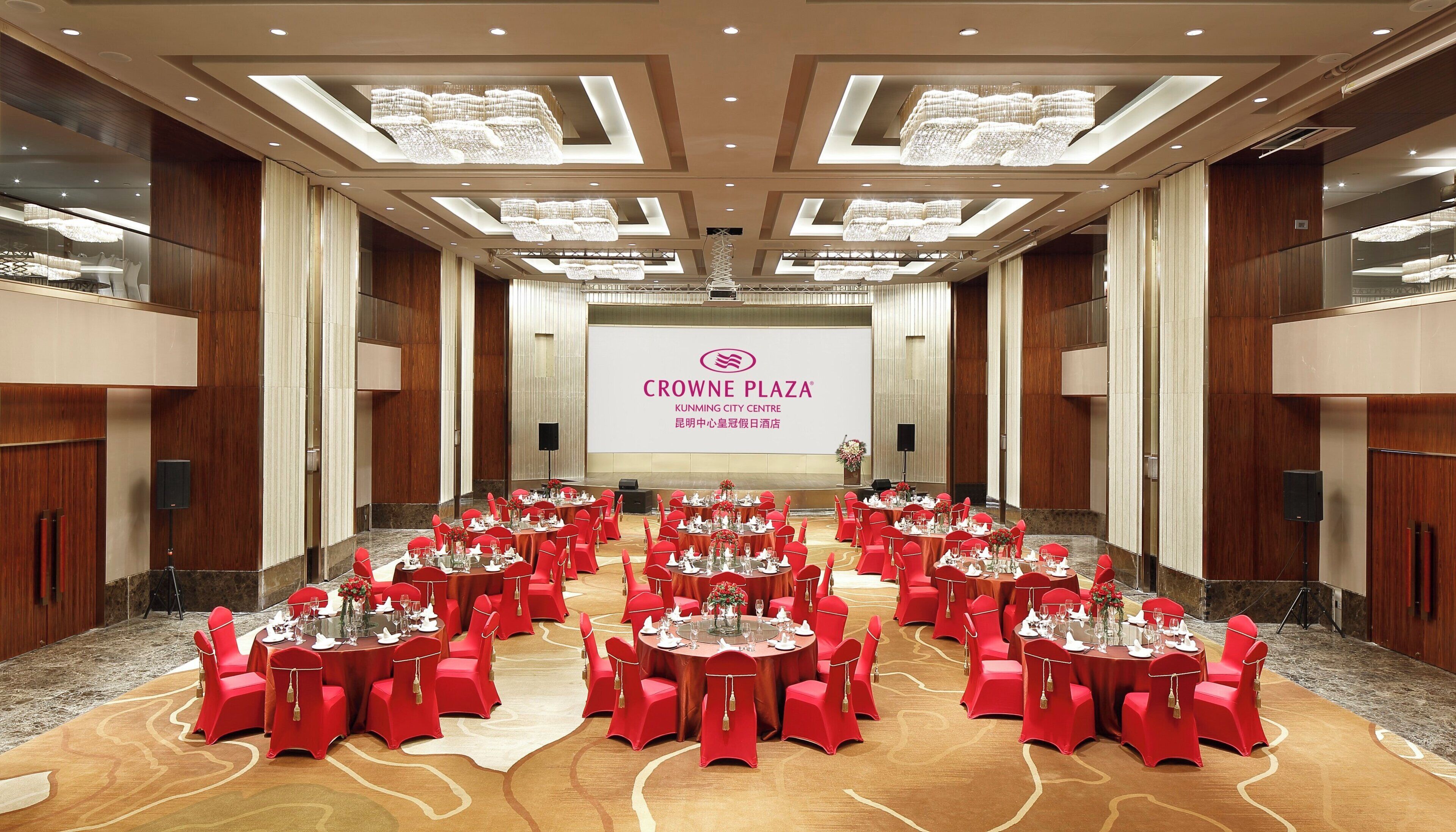 Un salón de conferencias con mesas redondas y sillas rojas, una pantalla grande que muestra el logotipo de Crowne Plaza y un panel de pared de madera.