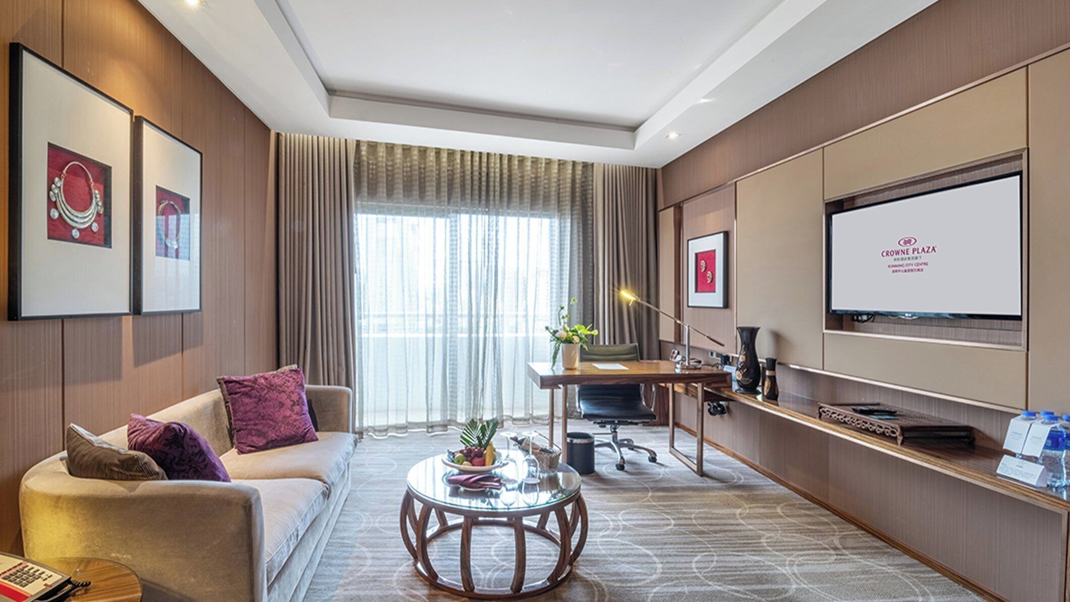 A hotel room with a sofa, a coffee table, a desk, a television, and a window with curtains.